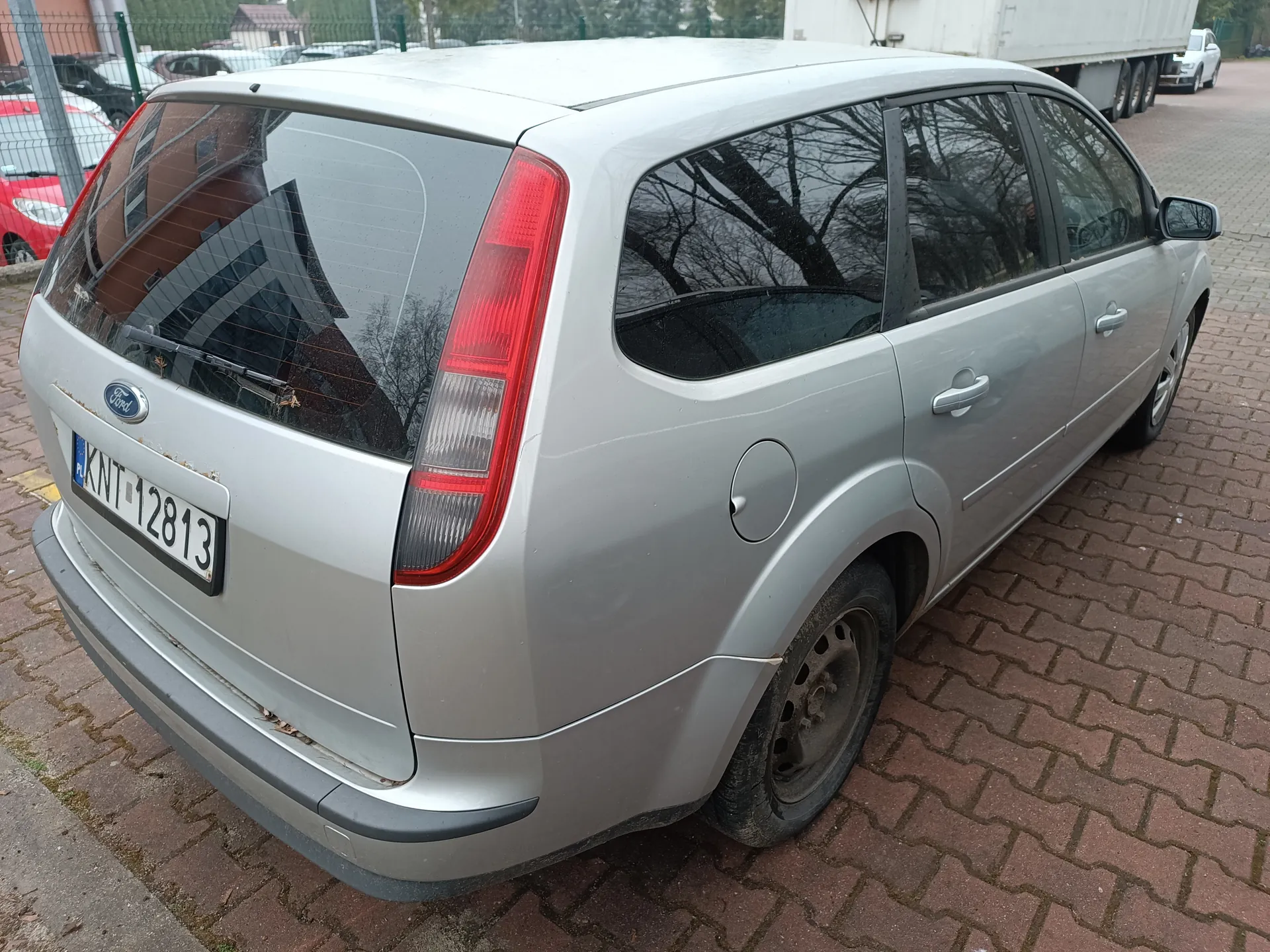 Srebrny samochód marki Ford, widok z tyłu, zaparkowany na ceglanym placu parkingowym. Tablica rejestracyjna KNT-12813 z kodem PL. W tle widoczne inne pojazdy, ogrodzenie i drzewa bez liści.