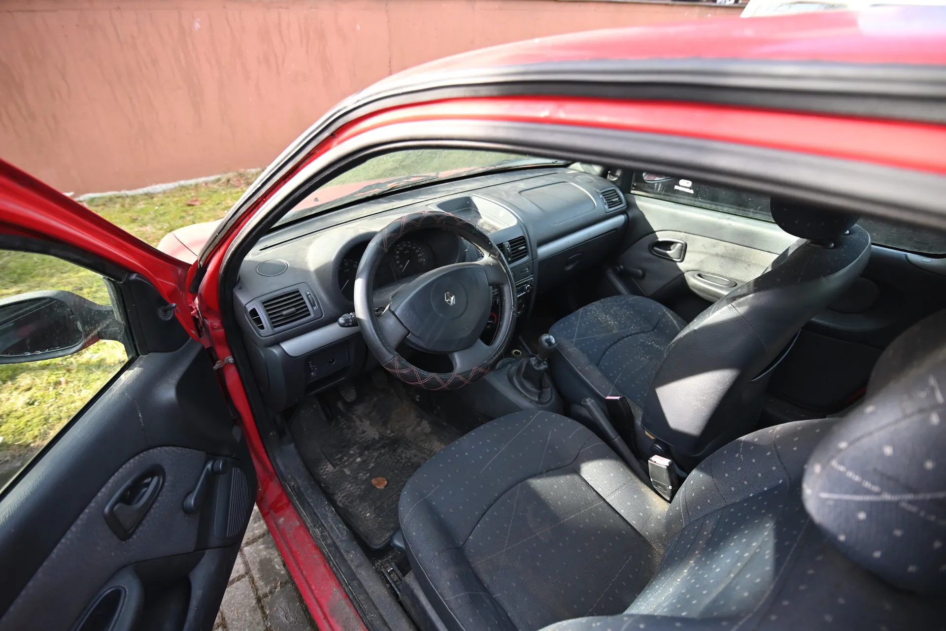 Wnętrze czerwonego samochodu z otwartą przodem drzwiami kierowcy. Widoczna czarna kierownica z logo Renault, czarne siedzenia z drobnym wzorem, szara deska rozdzielcza z wentylacjami i wskaźnikami. W tle brązowa ściana i trawa. Podłoga w aucie jest zabrudzona, a na siedzeniach widoczne są ślady zużycia.