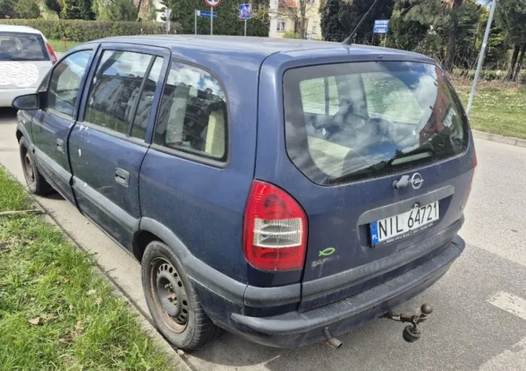 Niebieski samochód Opel typu kombi zaparkowany na poboczu drogi, widok od tyłu. Widoczna tablica rejestracyjna NIL 64721, czerwone światła tylnego, zaczep holowniczy oraz zabrudzona karoseria z oznakami rdzy na felgach. W tle trawa, biały samochód, drzewa, budynki oraz znaki drogowe. Horyzont i tekst na tablicy są poziome.