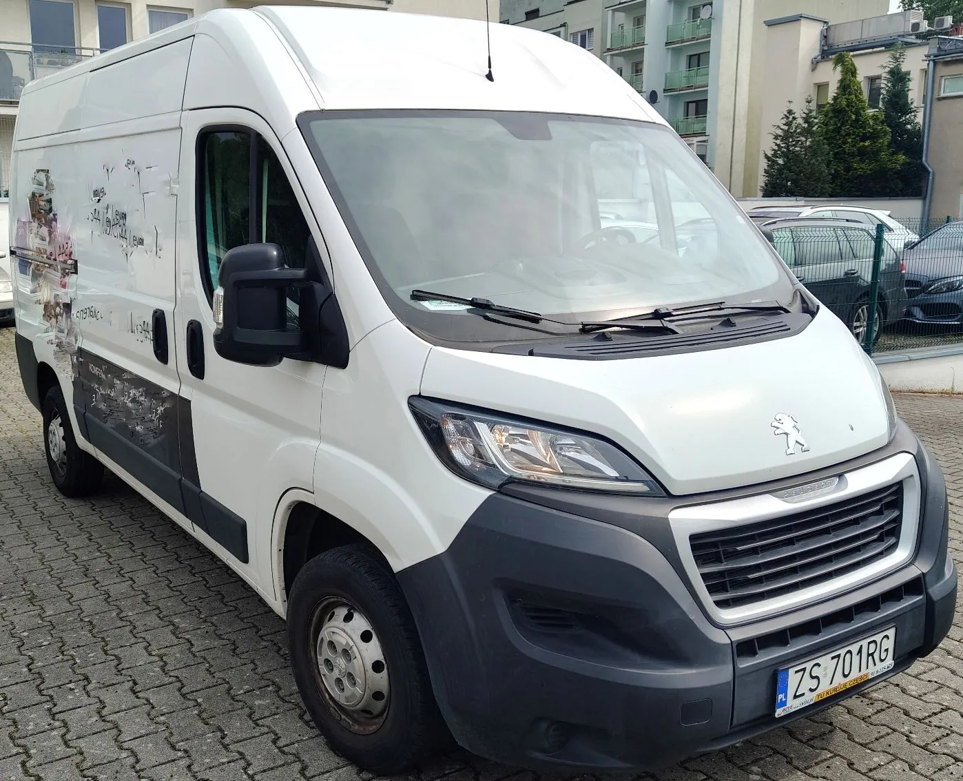 Biały furgon marki Peugeot zaparkowany na kostce brukowej. Pojazd ma czarny zderzak przedni oraz czarne lusterka boczne. Na boku vana widoczne są pozostałości po usuniętej oklejce reklamowej. Tablica rejestracyjna samochodu to ZS 701RG. W tle widać budynki mieszkalne oraz inne zaparkowane samochody.