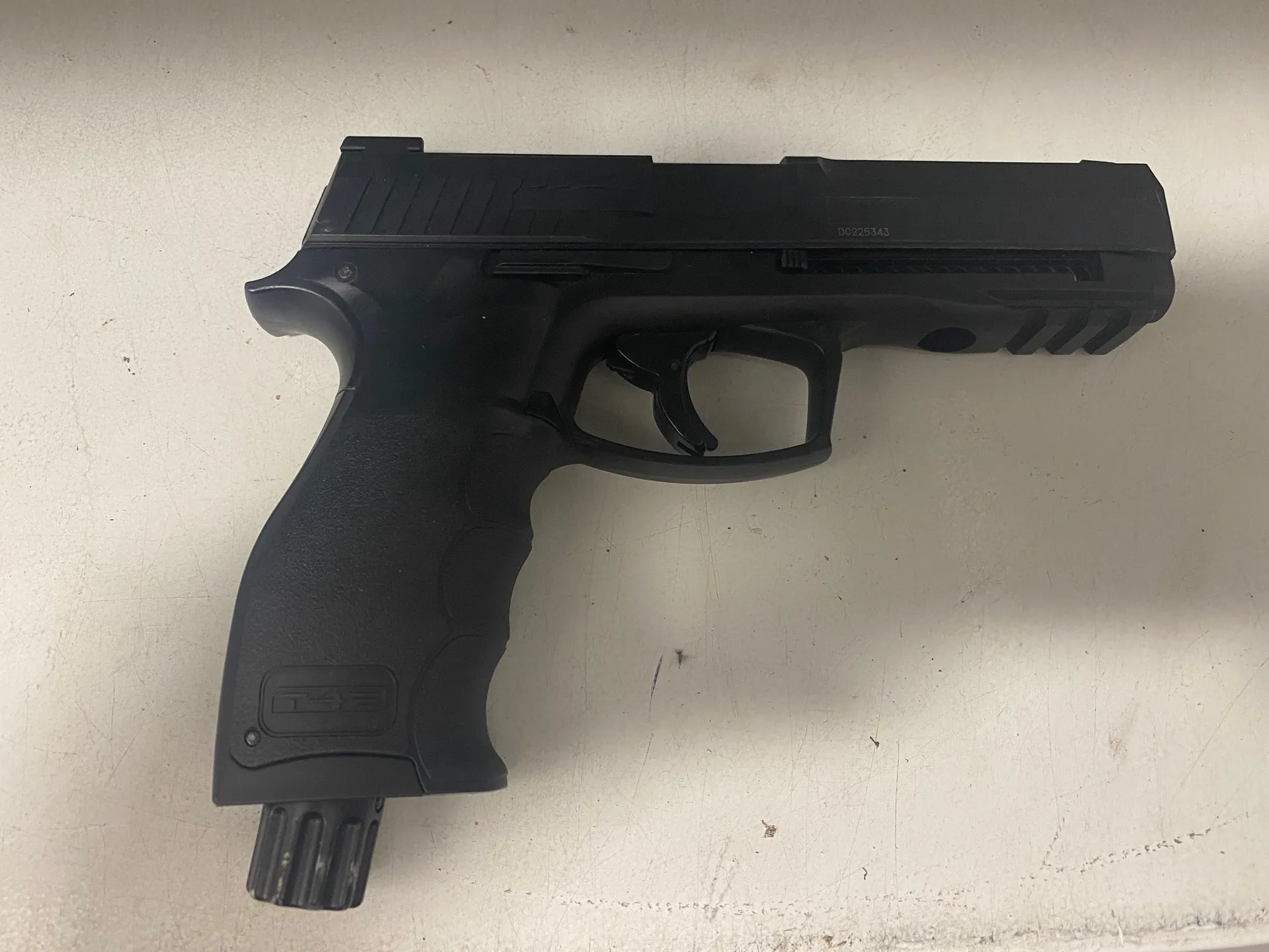 Czarny pistolet z logo T4E na rękojeści, cylindrycznym uzupełnieniem na lufie oraz widocznym numerem seryjnym DC225343 na zamku. Broń umieszczona na jasnej, zadrapanej powierzchni.