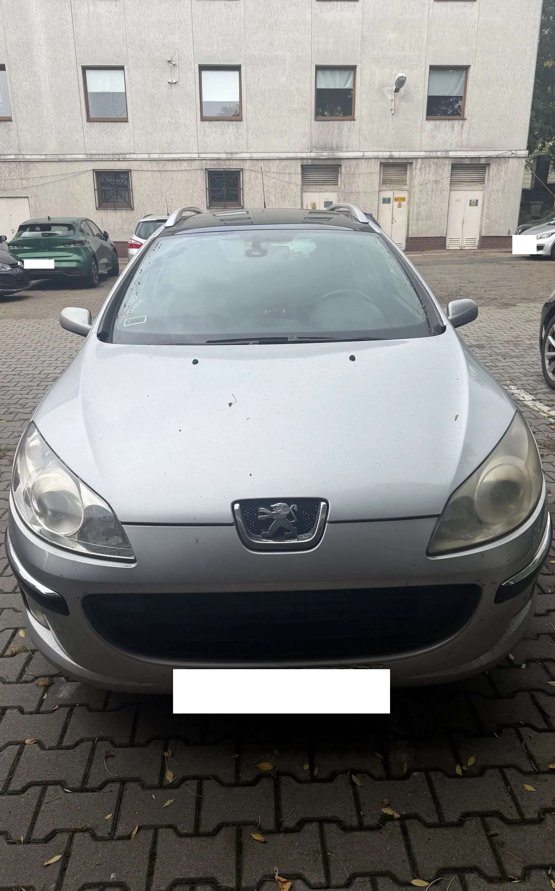 Srebrny samochód marki Peugeot zaparkowany przodem do obiektywu na kostce brukowej. W tle widoczny jest jasny, wielopiętrowy budynek z rzędem okien oraz inne zaparkowane samochody. Tablica rejestracyjna pojazdu jest zakryta białym prostokątem.