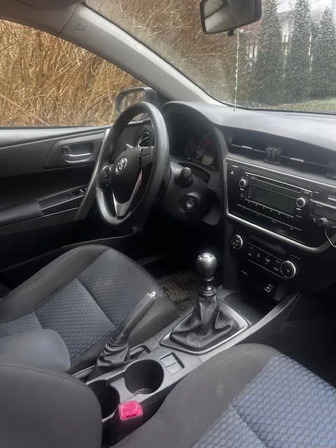 Wnętrze samochodu z widocznym kierownicą z logo, deską rozdzielczą z radiem i przyciskami, dźwignią zmiany biegów oraz czarnymi siedzeniami z wzorzystą tapicerką. Przez przednią szybę widać suchą trawę i drzewa poza pojazdem. W lusterku wstecznym widoczny fragment otoczenia.