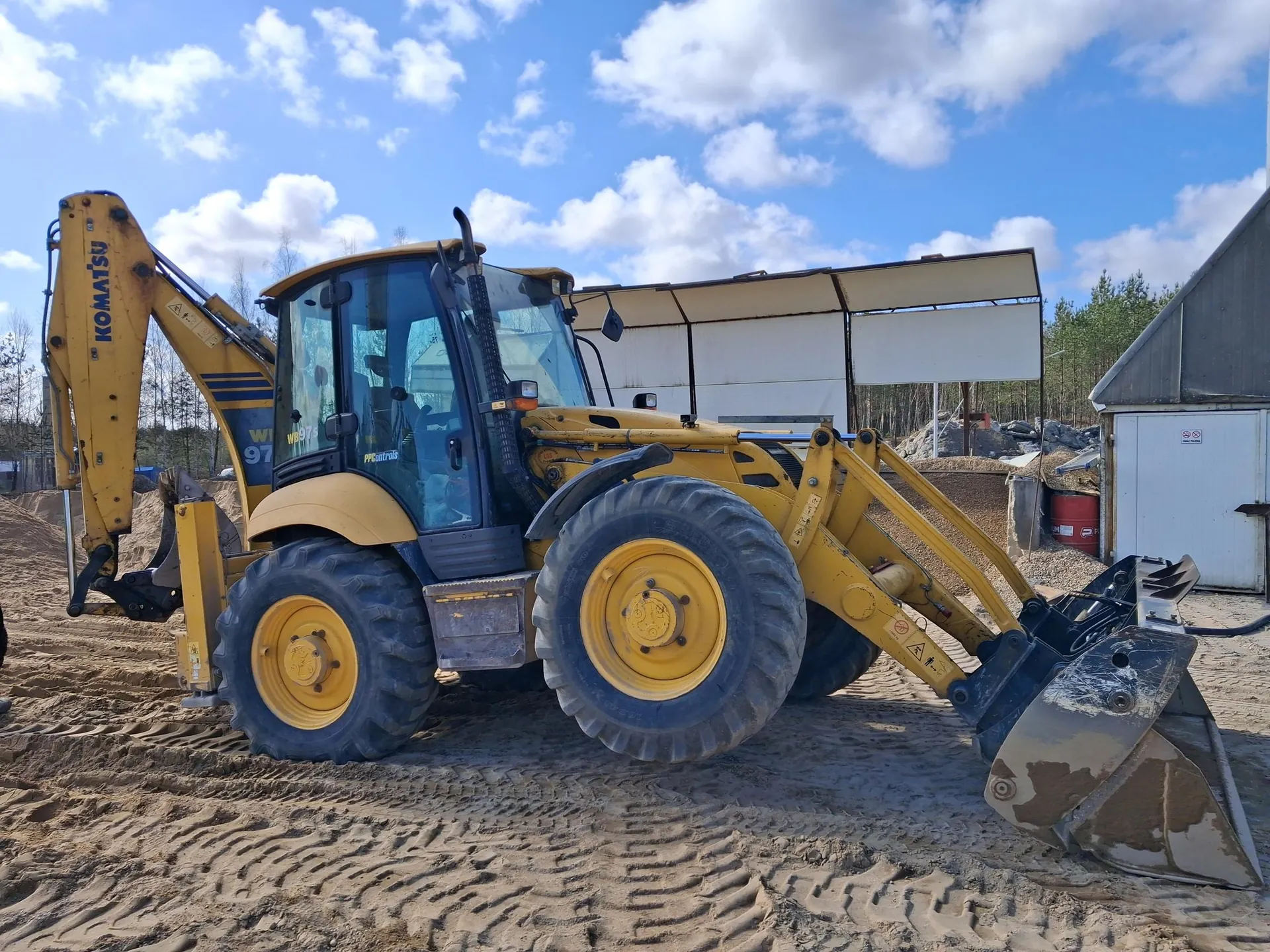 Żółty koparko-ładowarka marki Komatsu model WB97S stojąca na piaskowatej powierzchni z widocznymi śladami opon. Na ramieniu maszyny widnieje napis KOMATSU, a na boku kabiny model WB97S. W tle budynek z białymi drzwiami, czerwony beczka, stosy materiału budowlanego oraz niebo z białymi chmurami. Maszyna ma duże opony z żółtymi felgami i czarną łopatę na przodzie.