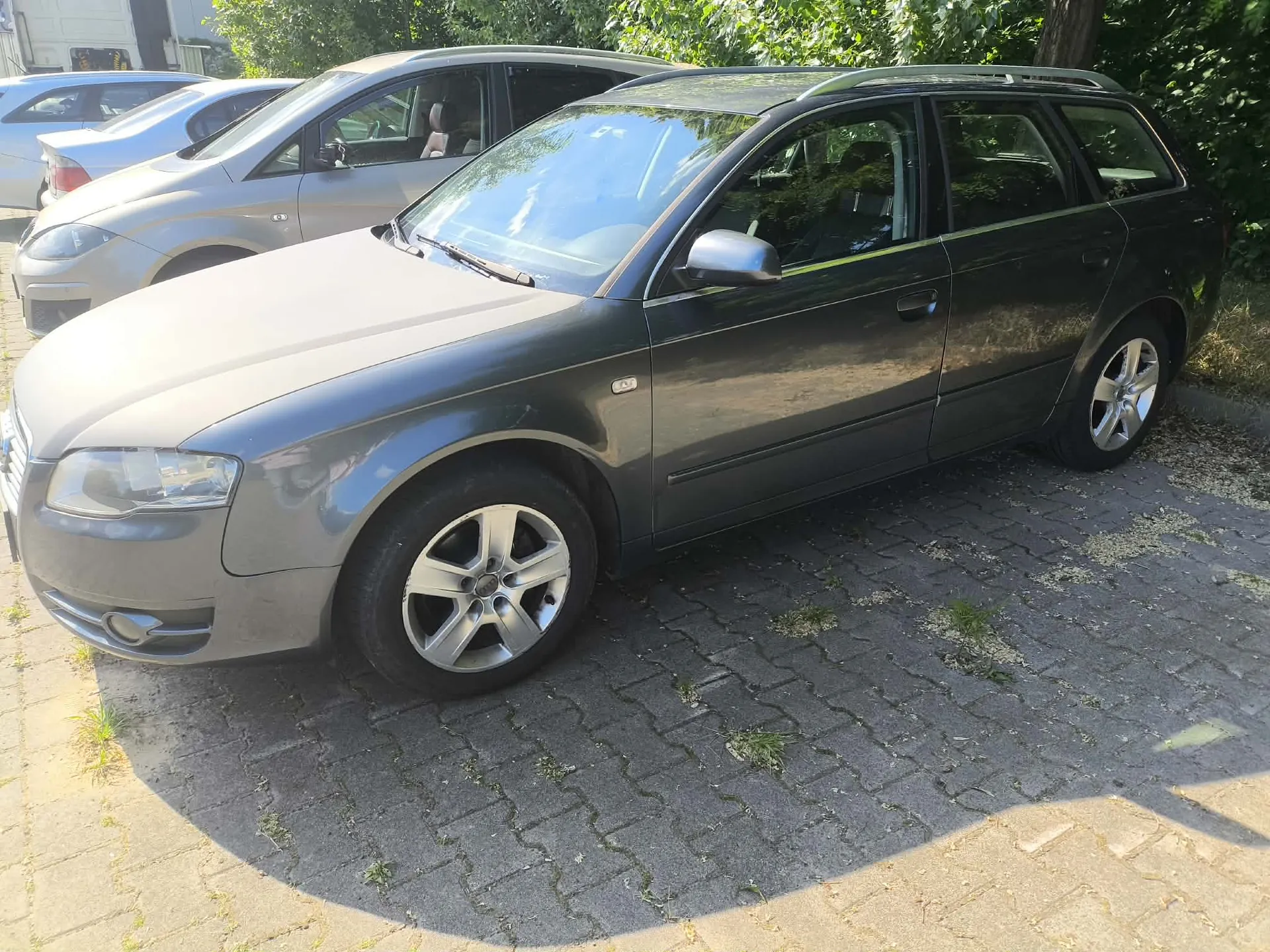 Szary samochód osobowy typu kombi zaparkowany na kostce brukowej. Pojazd jest widoczny z przodu i z boku. W tle widoczne są inne zaparkowane samochody oraz zielone drzewa.