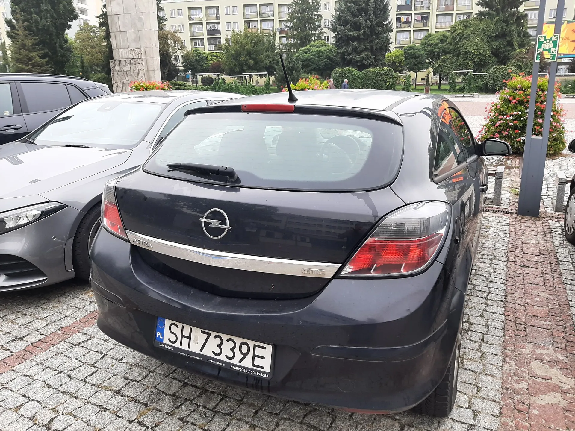 Tylny widok ciemnoszarego samochodu marki Opel Astra GTC zaparkowanego na kostce brukowej. Samochód ma polską tablicę rejestracyjną o numerze SH 7339E. Obok stoi srebrny samochód. W tle widoczne są bloki mieszkalne, zieleń oraz fragment pomnika.