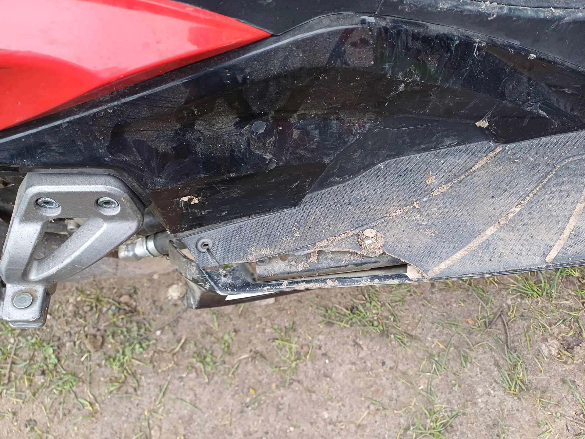 Zbliżenie na czerwono-czarną część pojazdu, prawdopodobnie motocykla. Srebrny metalowy uchwyt z dwiema śrubami, czarna powierzchnia z widocznymi zadrapaniami, brudem i śladami zużycia. Tło: ziemia z rzadką trawą. Brak czytelnych napisów.