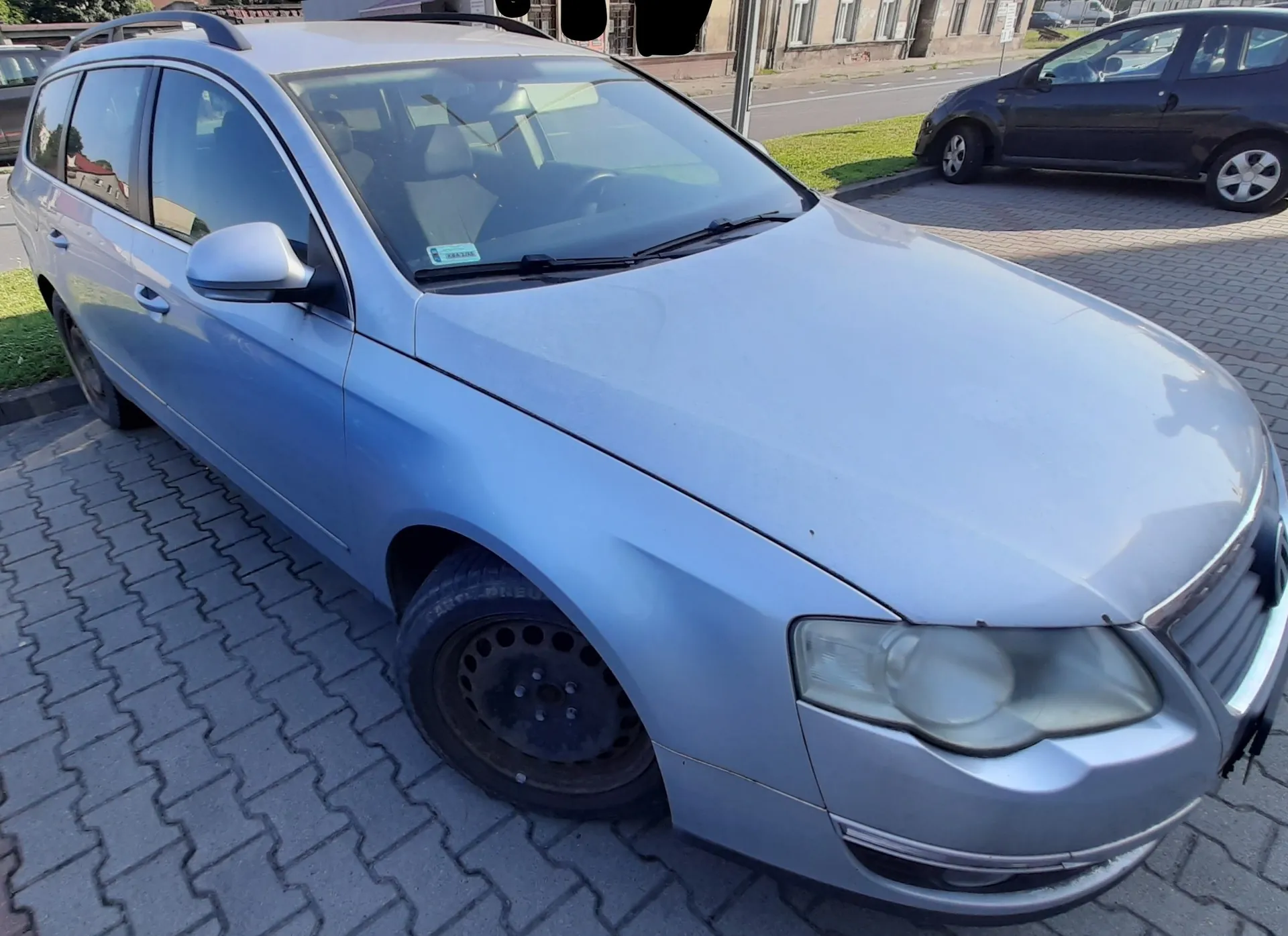 Srebrny samochód osobowy typu kombi zaparkowany na kostce brukowej. Widok z przodu i z boku pojazdu. W tle widać fragmenty budynków oraz inny, czarny samochód zaparkowany obok.