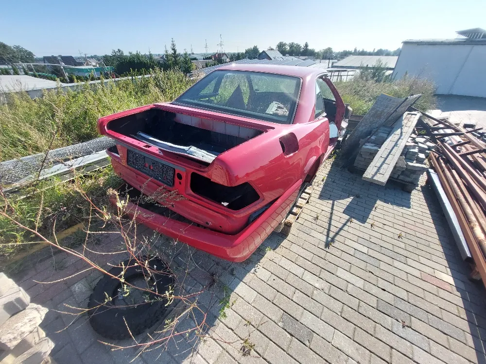 Czerwony samochód typu coupe z usuniętą tylną szybą i klapą bagażnika, stojący na kostce brukowej. Pojazd jest w złym stanie technicznym, z widocznymi brakami w elementach nadwozia z tyłu. Obok samochodu leżą drewniane deski, metalowe profile oraz stara opona. W tle widoczne są niskie budynki, roślinność i bezchmurne niebo.