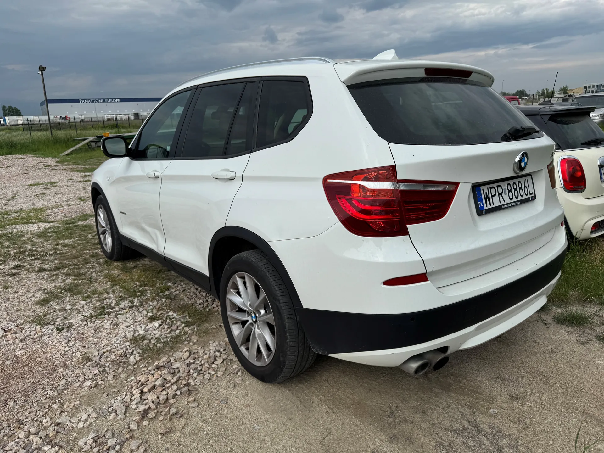 Biały SUV marki BMW z widocznym tyłem, tablicą rejestracyjną WPR 8886L z kodem PL oraz logo BMW. Po prawej stronie częściowo widoczny biały samochód. Tło: budynek z napisem PANATTONI EUROPE, niebo z chmurami, podłoga z żwiru i trawy.