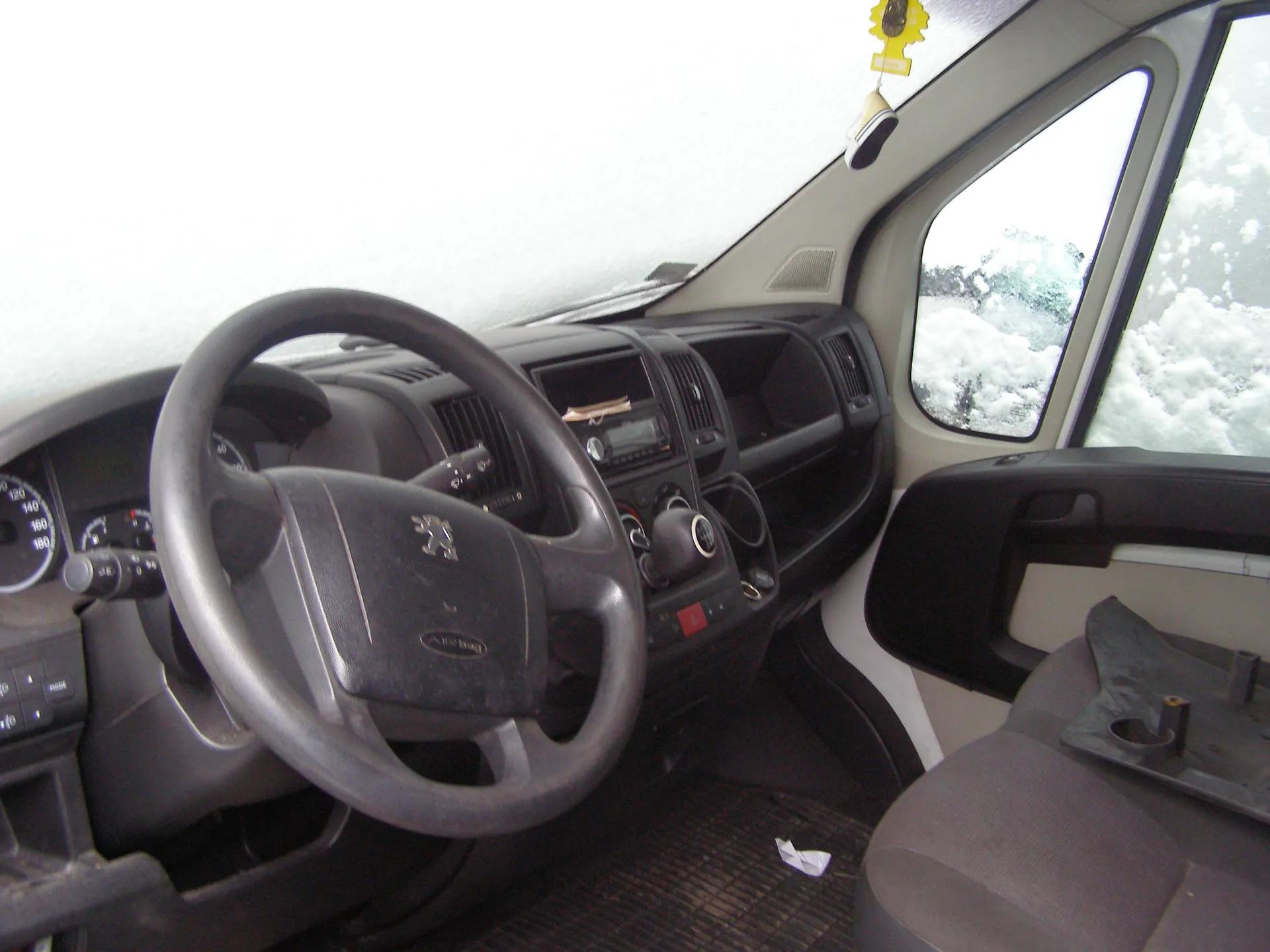 Wnętrze samochodu Peugeot z kierownicą z logo i napisem "Airbag". Przednią szybę i okna pokrywa śnieg. Deska rozdzielcza z licznikami (0–180 km/h), odtwarzaczem i przyciskami. Wisi żółty odświeżacz. Na siedzeniu pasażera przedmioty, a na podłodze maty i biała kartka.