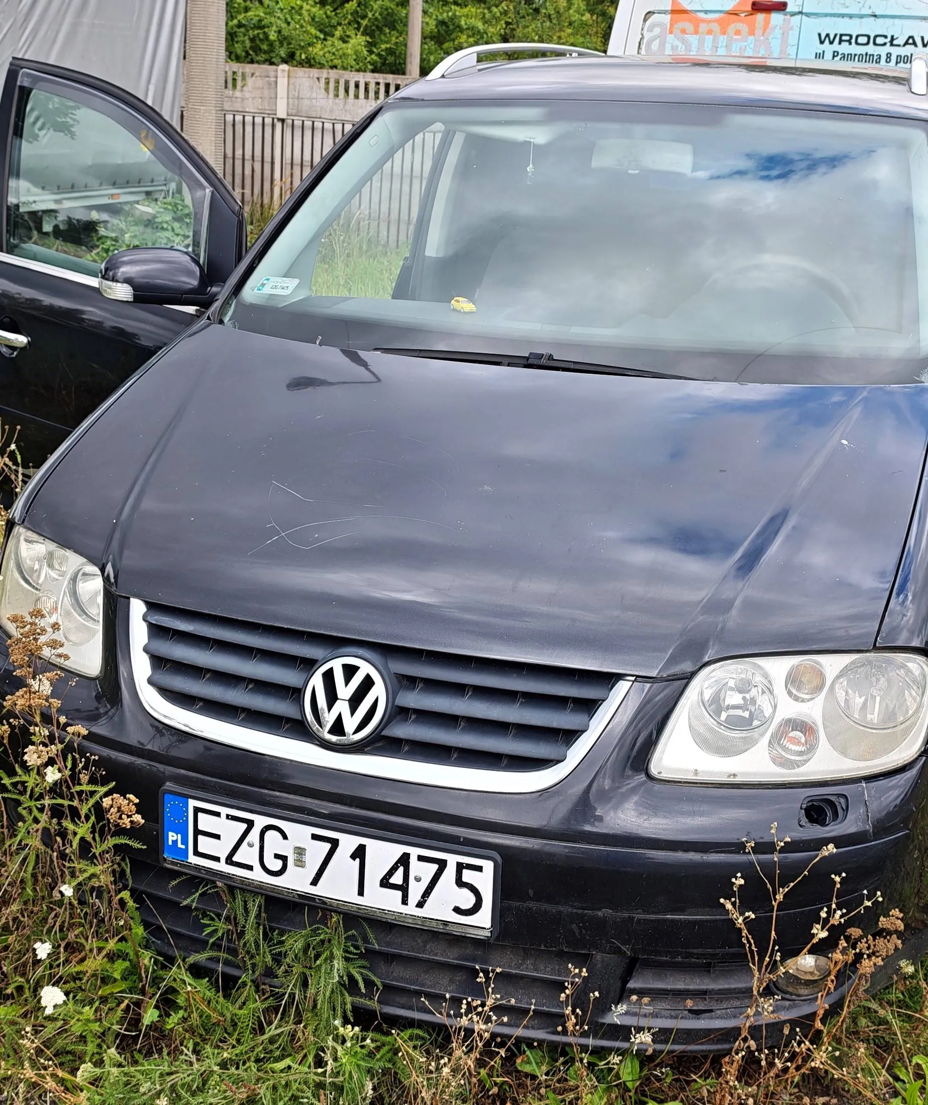 Przód ciemnogranatowego samochodu marki Volkswagen z widocznym logo na grillu. Pojazd posiada polską tablicę rejestracyjną o numerze EZG 71475. Samochód stoi na trawie, a w tle widoczny jest fragment białego ogrodzenia oraz niebieski szyld z napisem 'WROCŁAW ul. Panrotna 8 pol'. Lewe przednie drzwi samochodu są otwarte.