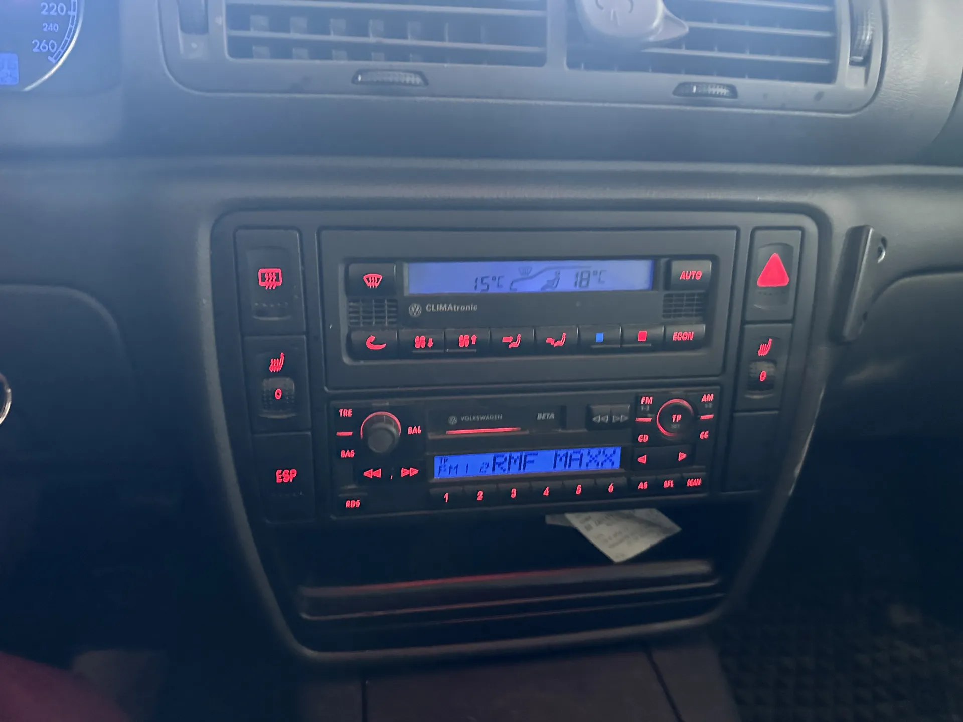Deska rozdzielcza samochodu z systemem klimatyzacji CLIMATronic i radiem. Wyświetlacz klimatyzacji pokazuje 15°C i 18°C. Radio marki Volkswagen z oznaczeniem BETA, na wyświetlaczu widoczny napis 'FM 1 2 RMF MAXXX'. Czerwone przyciski sterujące z symbolami (m.in. ESP, AUTO, ECON), wyłącznik awaryjny z czerwonym trójkątem. W dolnej części znajduje się bilet lub dokument pod radiem. W tle widoczny fragment wskaźnika prędkości z niebieskim oświetleniem.