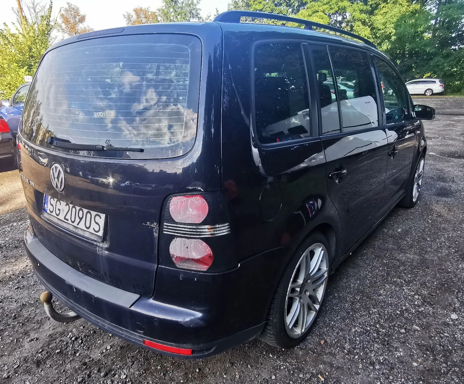 Tylny widok ciemnogranatowego samochodu marki Volkswagen w nadwoziu kombi, zaparkowanego na żwirowym podłożu. Pojazd posiada tablicę rejestracyjną o numerze SG 2090S oraz hak holowniczy. W tle widoczne są zielone drzewa i fragmenty innych samochodów.