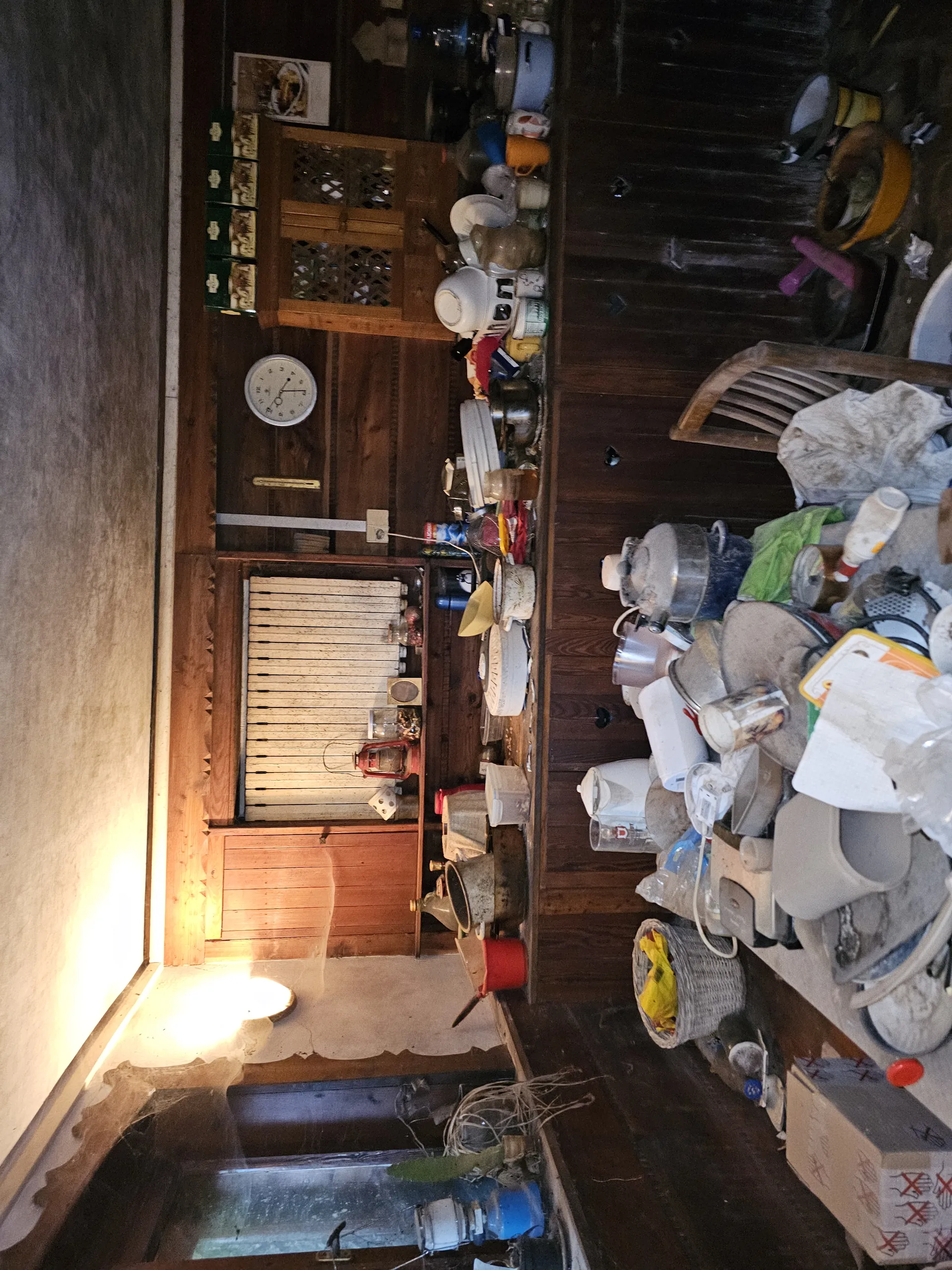 Wnętrze pomieszczenia z drewnianymi ścianami, wypełnione różnymi przedmiotami. Na półkach i powierzchniach znajdują się naczynia kuchenne, miski, kubki, garnki, plastikowe pojemniki oraz inne artykuły. Widać zegar ścienny z białą tarczą, drewnianą szafkę z ozdobnymi otworami, pudełka i krzesło. Podłoga betonowa, po prawej stronie okno z oświetleniem wpadającym do wnętrza. Przedmioty są gęsto rozmieszczone na różnych powierzchniach.
