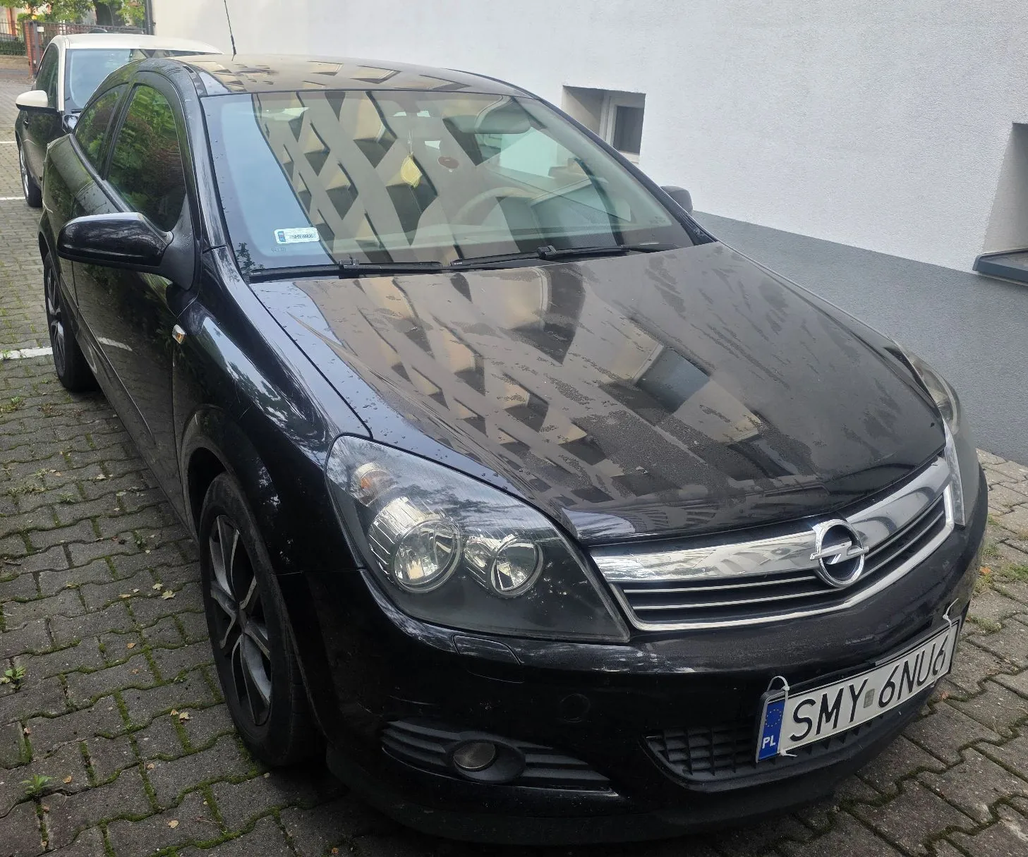 Czarny samochód osobowy marki Opel zaparkowany na kostce brukowej przed jasnym budynkiem. Widoczna jest przednia część pojazdu z tablicą rejestracyjną o numerze SMY 6NU6.