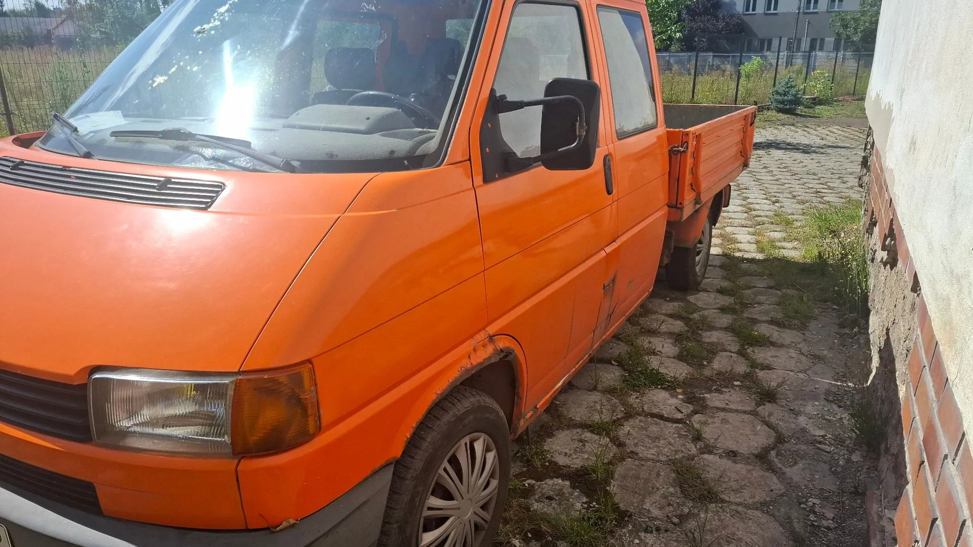 Pomarańczowy samochód dostawczy typu pick-up zaparkowany na podjeździe z kostki brukowej obok białej ściany budynku. Widoczna jest przednia i boczna część pojazdu z otwartą przestrzenią ładunkową z tyłu.
