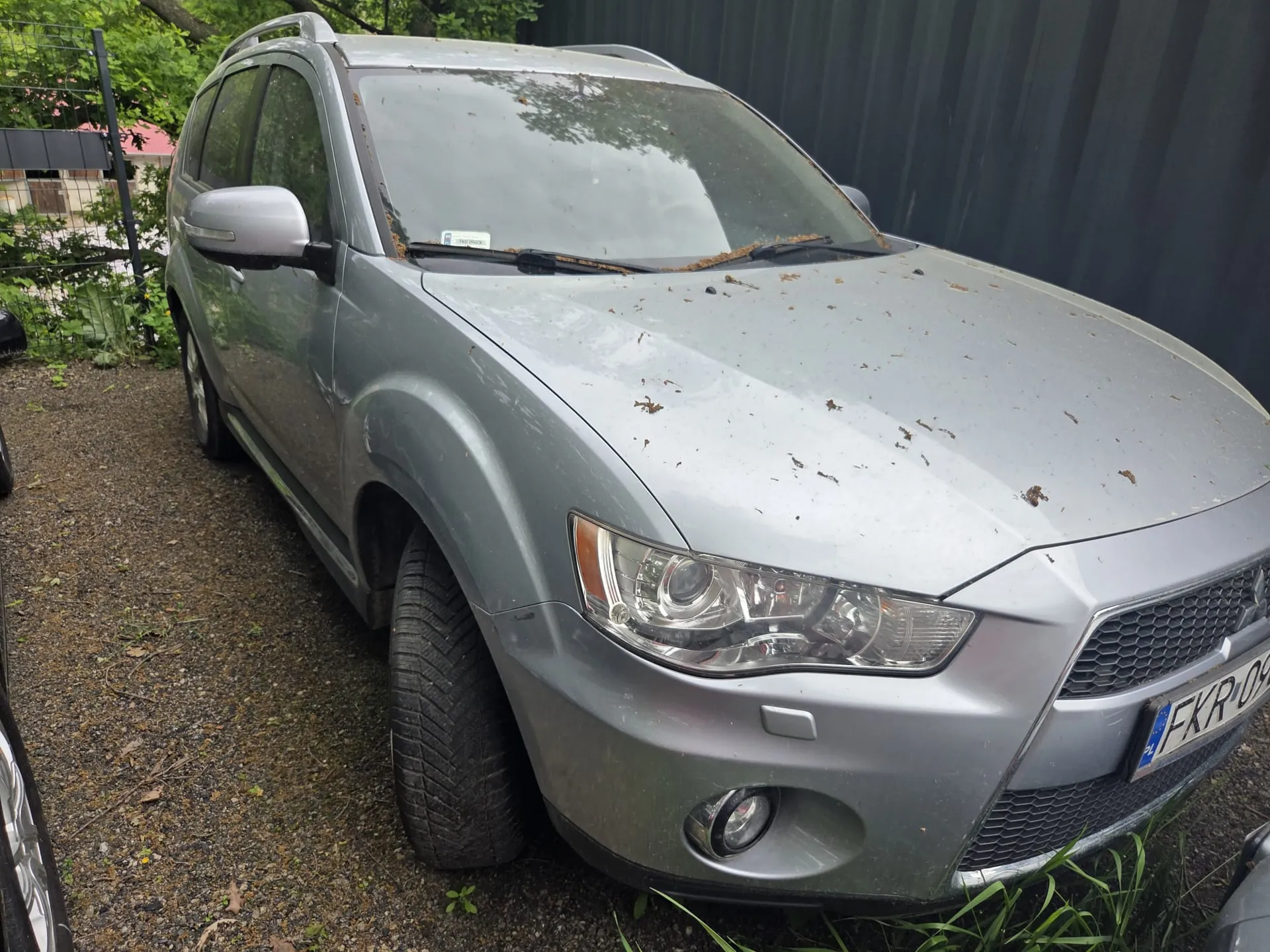 Srebrny samochód typu SUV zaparkowany na żwirowym podłożu. Na masce widoczne są brudne plamy i liście. Przedni reflektor i atrapa chłodnicy są wyraźne. Tablica rejestracyjna z napisem FKR-09 oraz oznaczeniem PL. W tle czarna metalowa bariera, zielone krzewy i fragment ogrodzenia. Po lewej stronie widoczna część innego pojazdu.