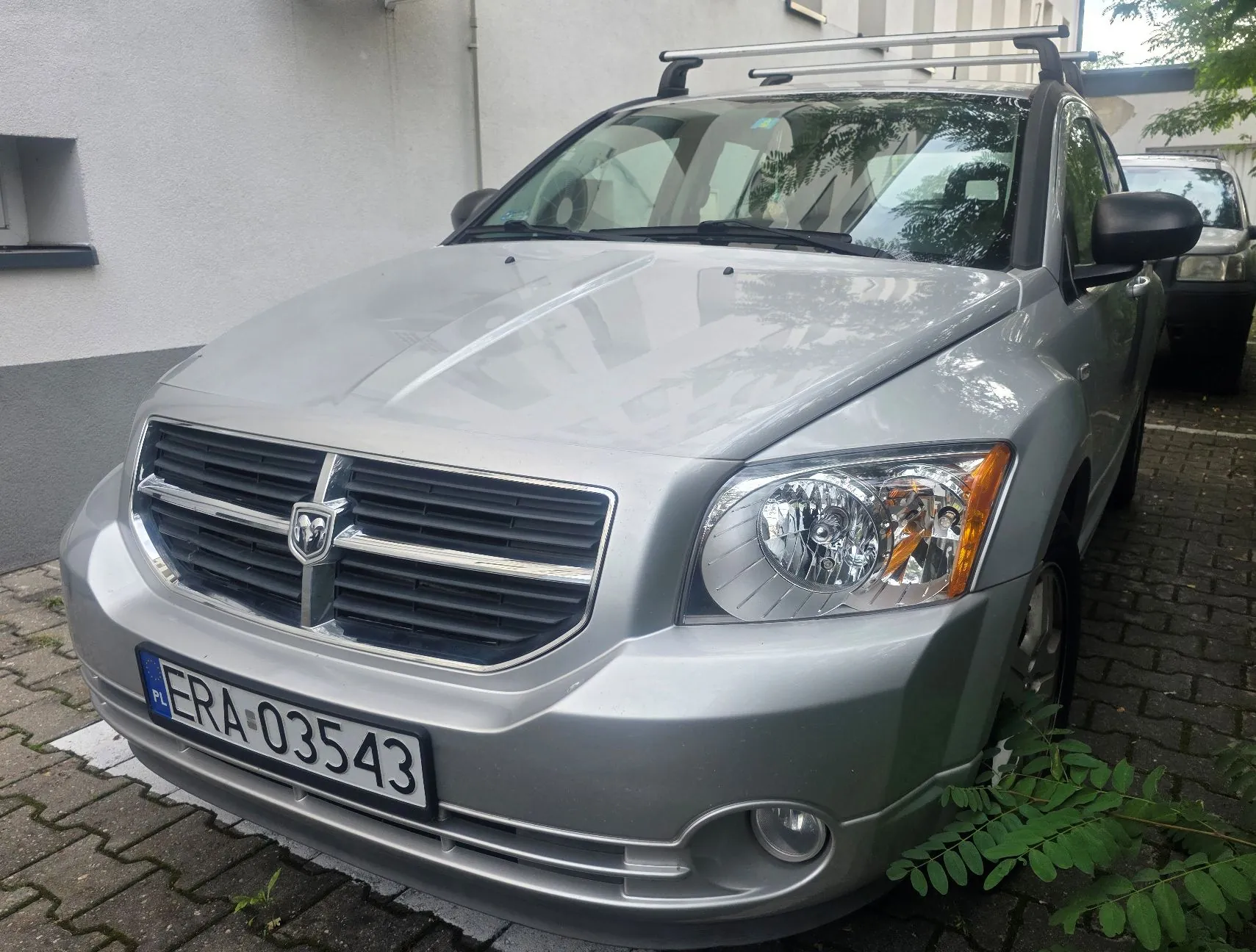 Srebrny samochód marki Dodge zaparkowany na kostce brukowej przed jasną ścianą budynku. Pojazd posiada zamontowany na dachu srebrny bagażnik dachowy. Na przednim zderzaku znajduje się polska tablica rejestracyjna o numerze ERA 03543. W prawym dolnym rogu widoczne są zielone liście rośliny.