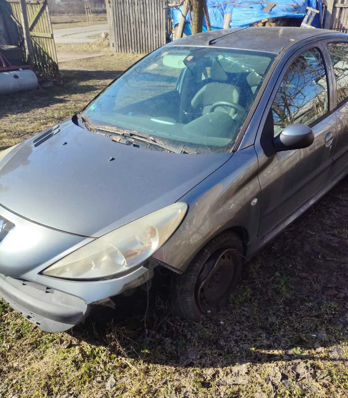 Zniszczony szary samochód osobowy zaparkowany na trawiastym i błotnistym terenie. Przednia część pojazdu jest uszkodzona: brakuje zderzaka, widoczne są deformacje maski oraz uszkodzone lewe przednie światło. W tle drewniane ogrodzenie, niebieskie tarpauliny przykrywające przedmioty oraz metalowa konstrukcja po lewej stronie. Samochód stoi na nierównym terenie z fragmentami trawy i liści.