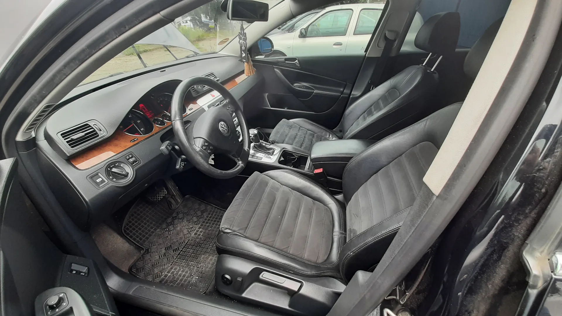Wnętrze samochodu marki Volkswagen z czarnymi skórzonymi fotelami, kierownicą z logo VW, deską rozdzielczą z drewnianą obwódką i licznikami. Widoczne są przyciski klimatyzacji, dźwignia zmiany biegów oraz dywaniki podłogowe. W lusterku wstecznym wiszą ozdoby. Poza samochodem, przez przednią szybę, widać inny pojazd i teren z trawą.