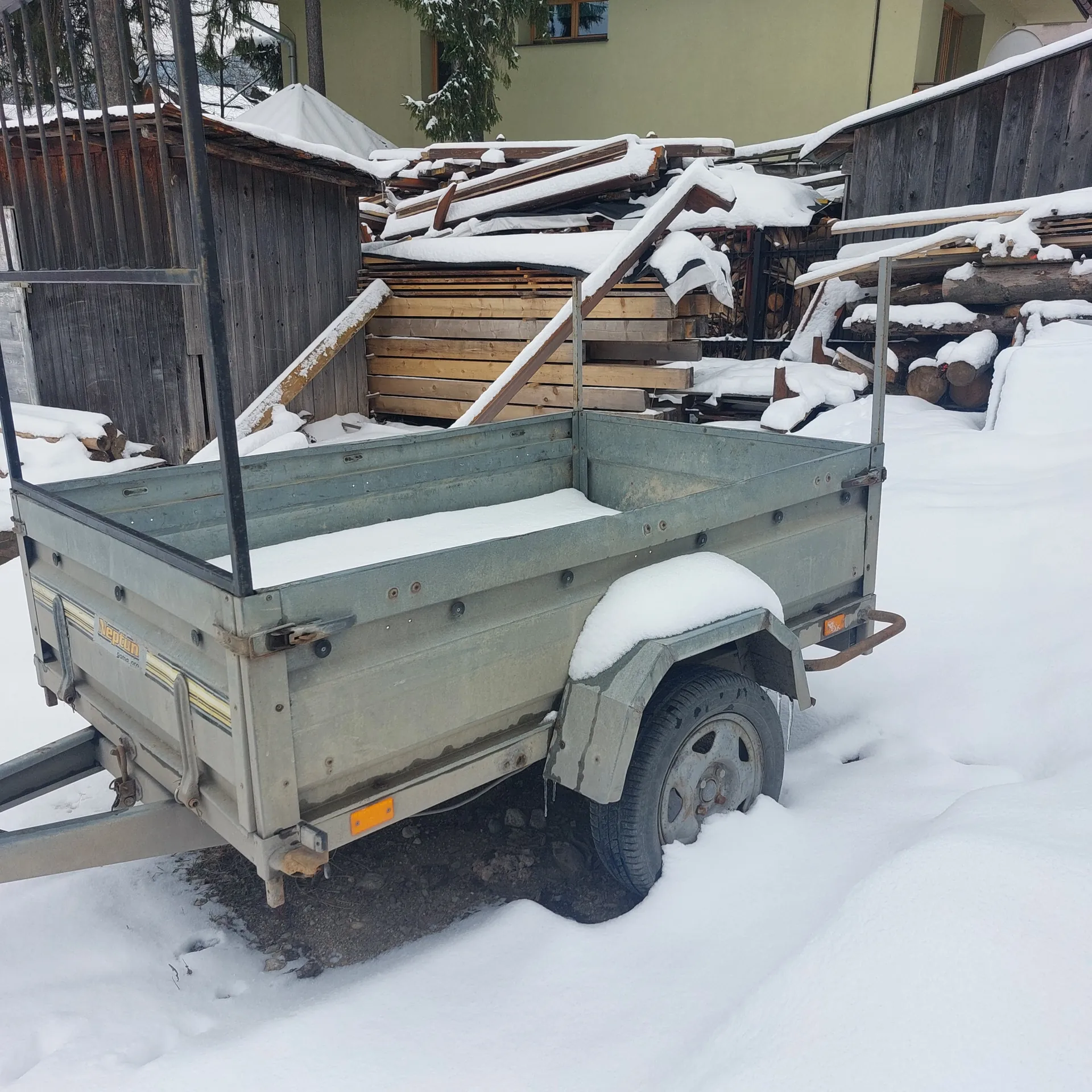 Zimowy krajobraz z metalową przyczepą pokrytą śniegiem. Przyczepa ma koło, pomarańczowe oznaczenia i częściowo widoczny napis na boku. Obok znajduje się drewniana konstrukcja z ułożonymi deskami i kłodami, również pokrytymi śniegiem. W tle widoczny jest budynek z jasnozieloną ścianą i oknem. Na lewo od przyczepy rośnie śniegiem pokryte drzewo oraz metalowe ogrodzenie. Śnieg pokrywa ziemię, w niektórych miejscach odsłaniając brudną ziemię i kamienie.