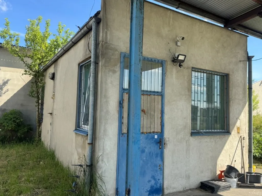 Zdjęcie narożnika niewielkiego, parterowego budynku o szarym, tynkowanym wykończeniu. W centralnej części znajduje się niebieskie, metalowe drzwi z kratą w górnej części. Po lewej stronie widoczne jest okno z kratami, a po prawej drugie, podobne okno. Nad drzwiami zamontowana jest kamera monitoringu oraz lampa zewnętrzna. Wzdłuż ściany biegnie rura spustowa. Po lewej stronie budynku rośnie zielone drzewo i trawa, a po prawej widać fragment zadaszenia oraz różne przedmioty gospodarcze na ziemi.