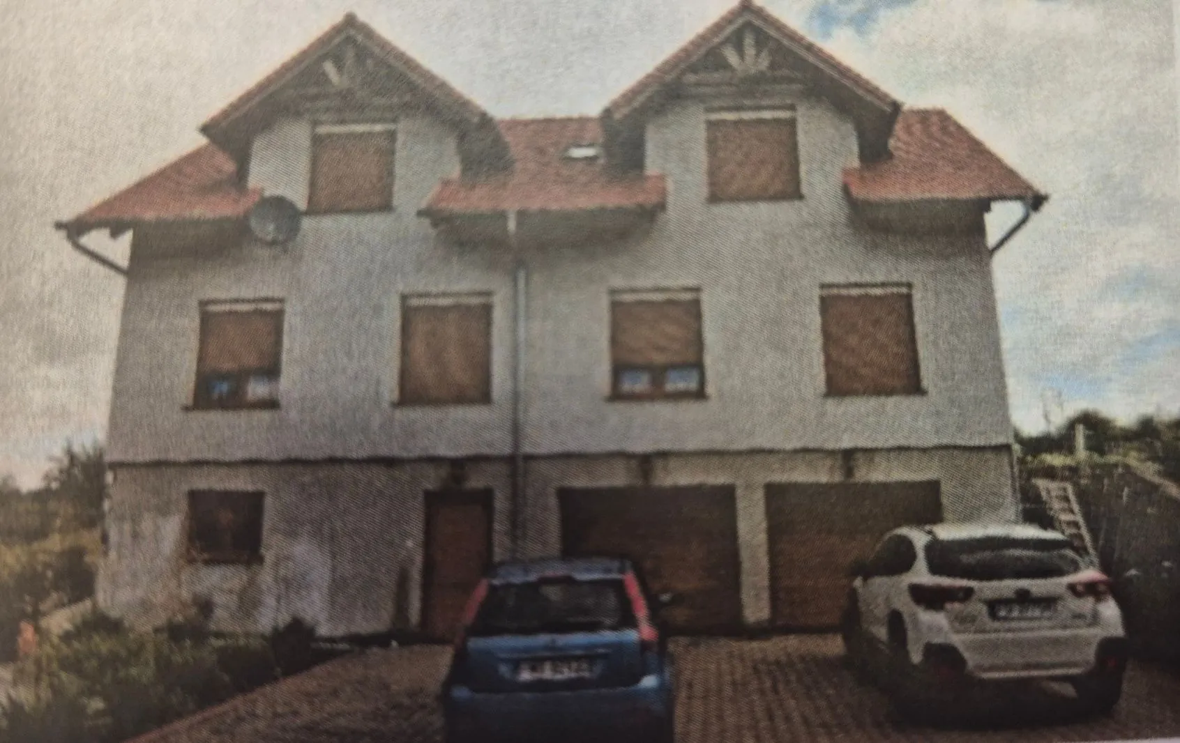 Zdjęcie dwukondygnacyjnego domu jednorodzinnego z poddaszem, wykończonego jasnym tynkiem i czerwonym dachem dwuspadowym. Wszystkie okna na piętrze i poddaszu są zasłonięte brązowymi roletami zewnętrznymi. Na parterze znajdują się dwa garaże oraz drzwi wejściowe. Przed domem zaparkowane są dwa samochody: niebieski hatchback z tyłu oraz biały samochód typu crossover po prawej stronie.