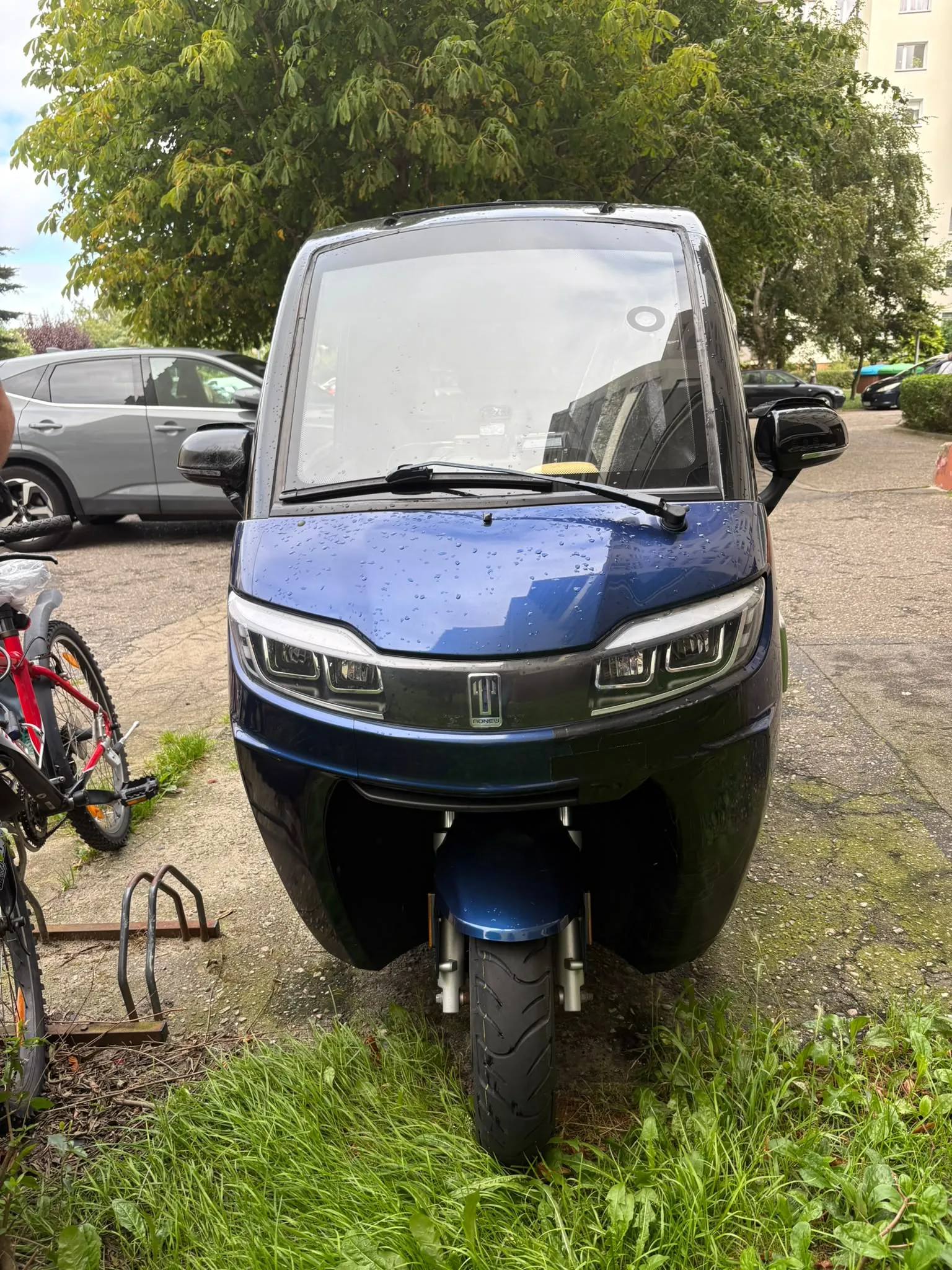 Niebieski, nowoczesny skuter elektryczny z zabudową w formie kabiny, zaparkowany na podjeździe. Pojazd ma dużą przednią szybę, dwa reflektory LED oraz lusterka boczne. W tle widoczny jest szary samochód osobowy, zielone drzewa oraz fragment budynku mieszkalnego. Po lewej stronie zdjęcia znajduje się fragment czerwono-czarnego roweru.