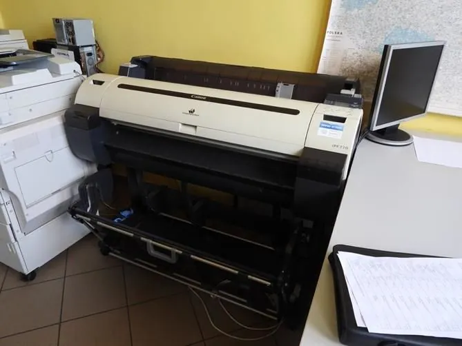 Duża, szeroka drukarka ploter marki Canon w kolorze białym i czarnym, stojąca w pomieszczeniu. Po lewej stronie znajduje się biała kserokopiarka, a po prawej monitor komputera na białym biurku. W tle widać żółtą ścianę i fragment mapy Polski.
