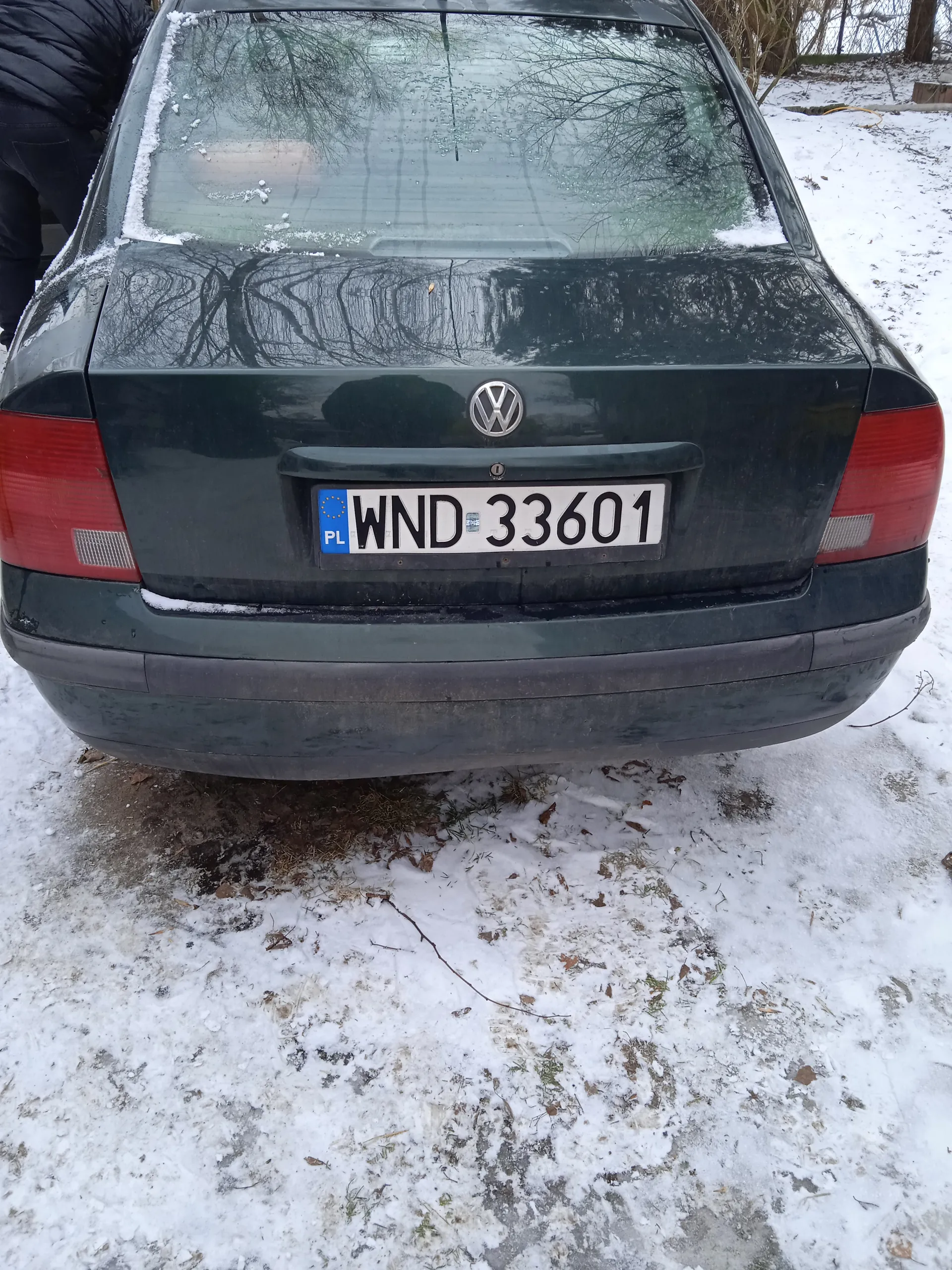 Tył ciemnozielonego samochodu marki Volkswagen z tablicą rejestracyjną WND 33601. Na lewej stronie tablicy widoczny niebieski pas z napisem PL i flagą Unii Europejskiej. Samochód stoi na śnieżnej powierzchni, na tle drzew i zimowego krajobrazu. Na szybie tylnej i bagażniku widoczne są ślady śniegu.
