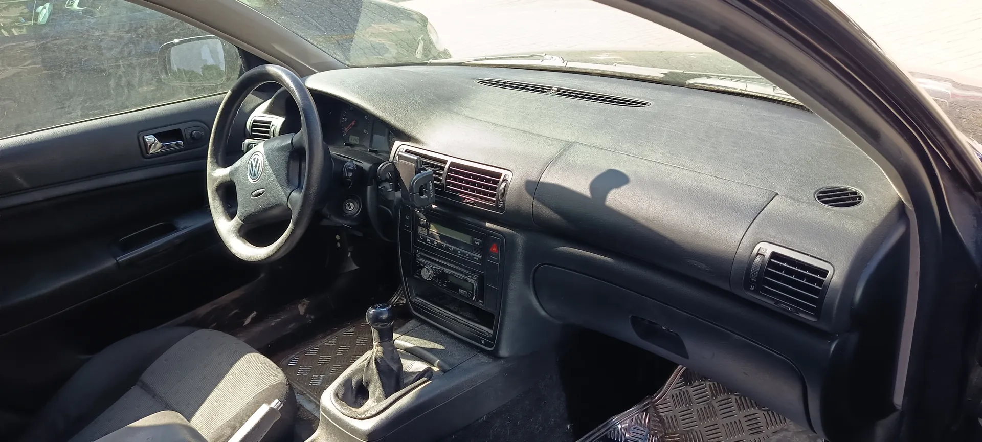 Wnętrze samochodu marki Volkswagen z widocznym kierownicą z logo VW, czarną deską rozdzielczą z wentylacjami, radioodtwarzaczem i licznikami. Przednie siedzenie pasażera oraz drzwi z uchwytem i lusterkiem bocznym. Na podłodze metalowe maty. Poza samochodem widoczne są inne pojazdy i asfaltowa powierzchnia.