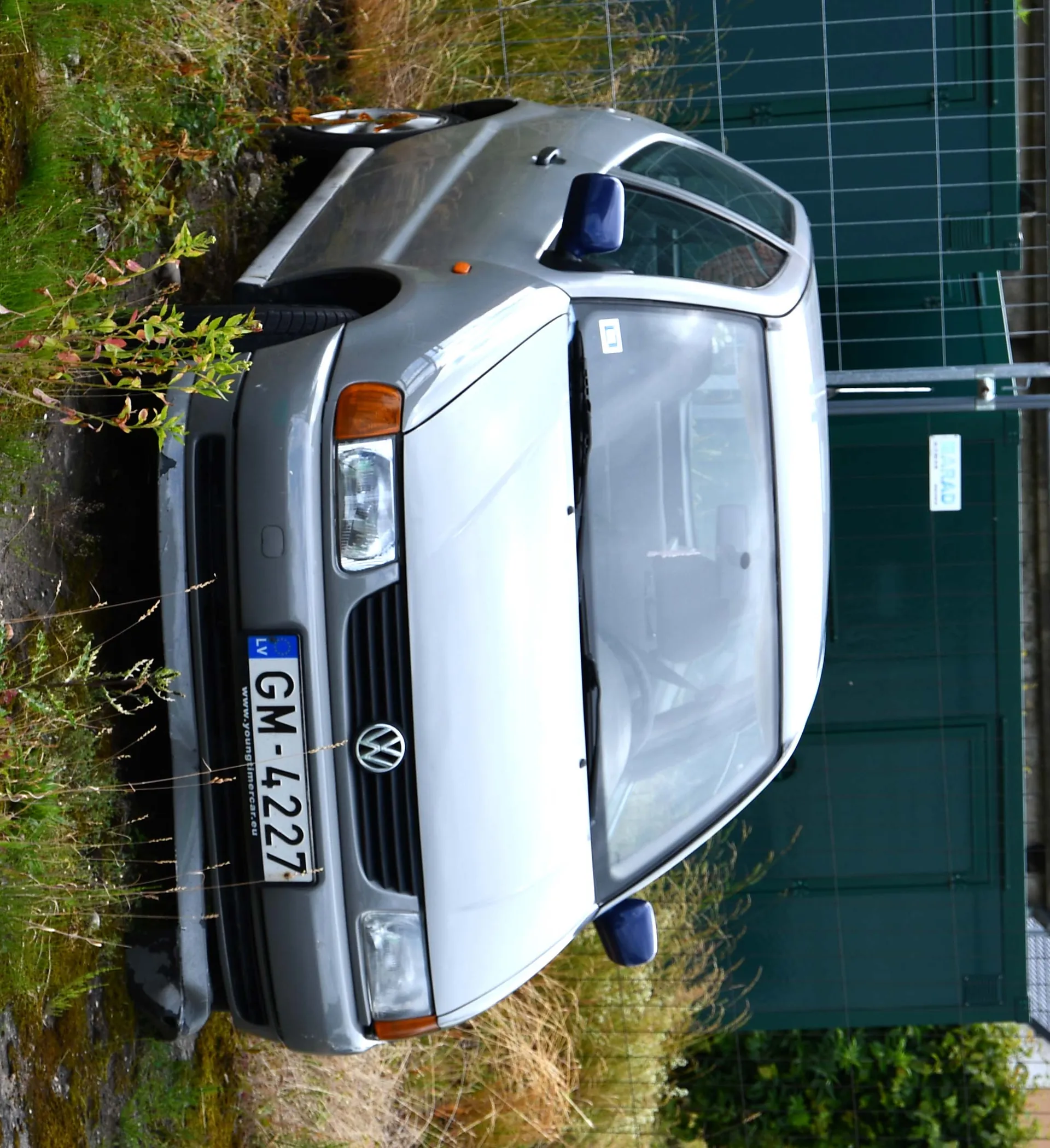 Srebrny samochód Volkswagen Polo z przodu, zaparkowany na trawie i mchu. Widoczna tablica rejestracyjna LV GM-4227 oraz adres www.youngtimer.eu pod nią. Tło stanowi zielone ogrodzenie z siatką i tablicą z napisem BARAD. Wokół rosną wysokie trawy i krzewy. Boczne lusterka samochodu są niebieskie, a na przedniej szybie znajduje się mały naklejony znaczek.