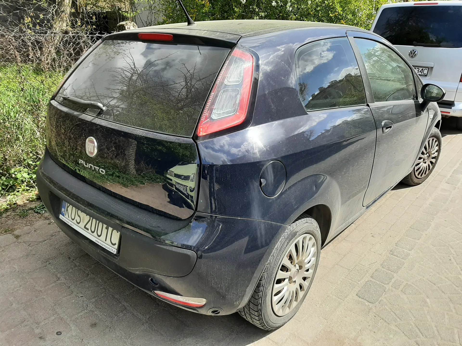 Ciemnoniebieski samochód Fiat Grande Punto zaparkowany tyłem do obserwatora. Widoczna tablica rejestracyjna KOS 2001C oraz napis "PUNTO" na bagażniku. Po prawej stronie częściowo widoczny srebrny samochód marki Volkswagen z tablicą 34C. Tło stanowią zielone krzewy, drzewa i brukowana powierzchnia. Odbicie nieba i otoczenia w szybach pojazdu.