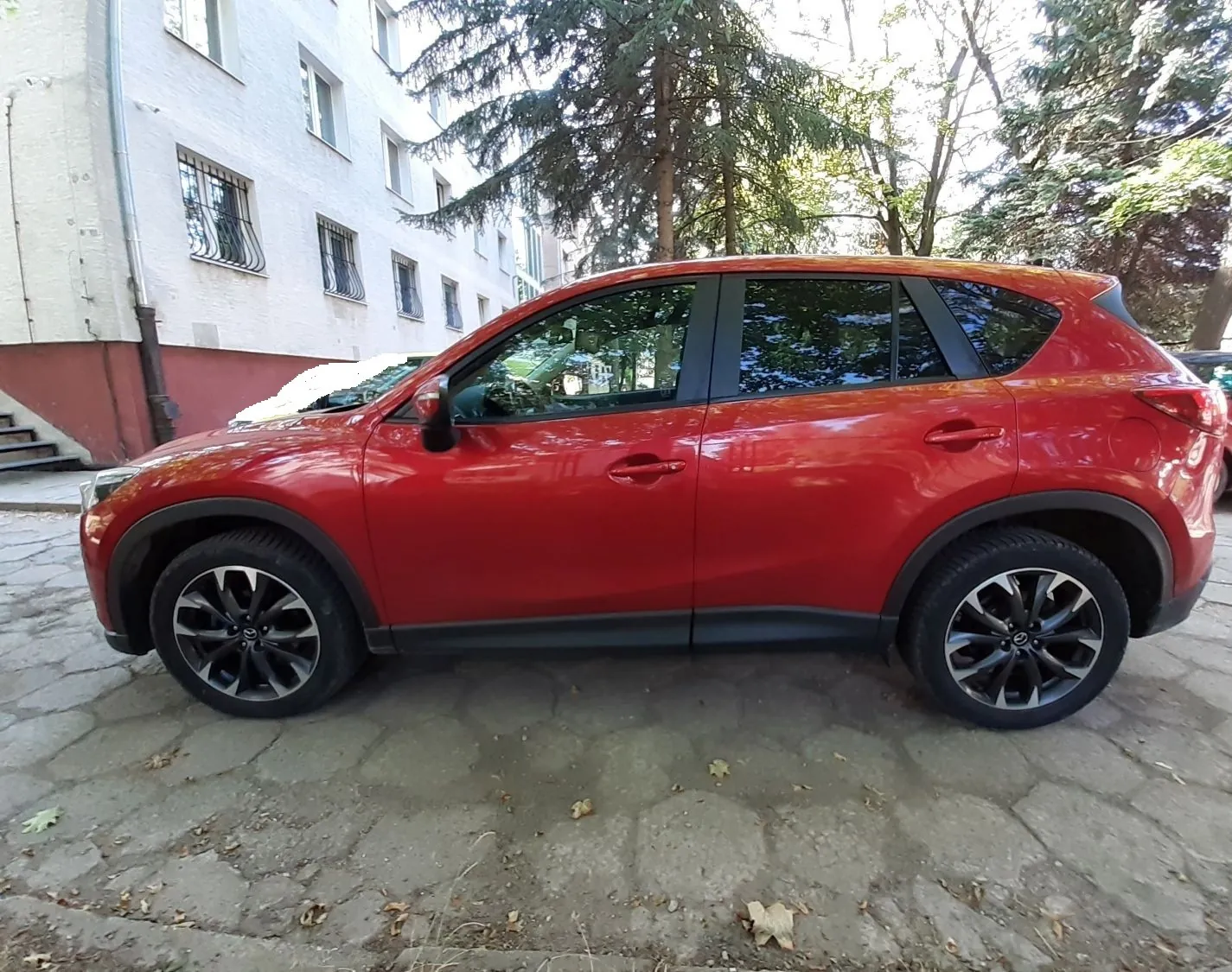 Czerwony samochód typu SUV zaparkowany bokiem na kostce brukowej. W tle znajduje się biały budynek mieszkalny z oknami oraz duże, zielone drzewa iglaste i liściaste.