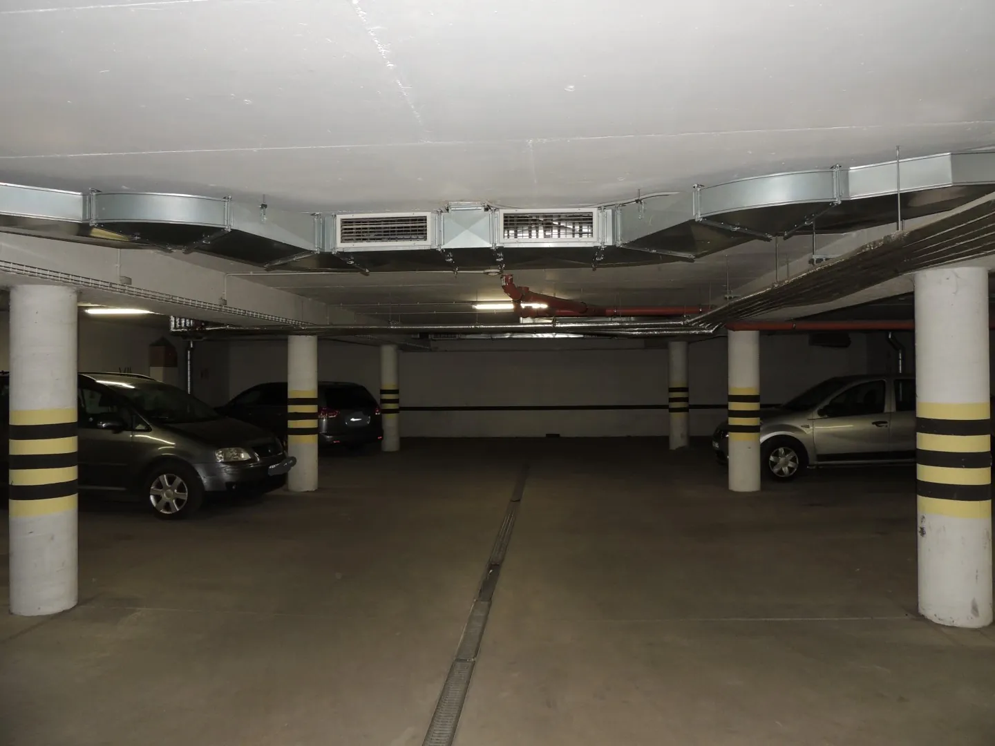 Podziemny parking z betonowym sufitem i podłogą. Na suficie zamontowane są srebrne kanały wentylacyjne. W przestrzeni znajdują się białe, okrągłe filary z poziomymi, czarnymi pasami. Zaparkowane są trzy samochody: szary samochód osobowy po lewej, ciemny samochód w głębi oraz srebrny samochód po prawej stronie. Przez środek podłogi przebiega metalowy odpływ liniowy.