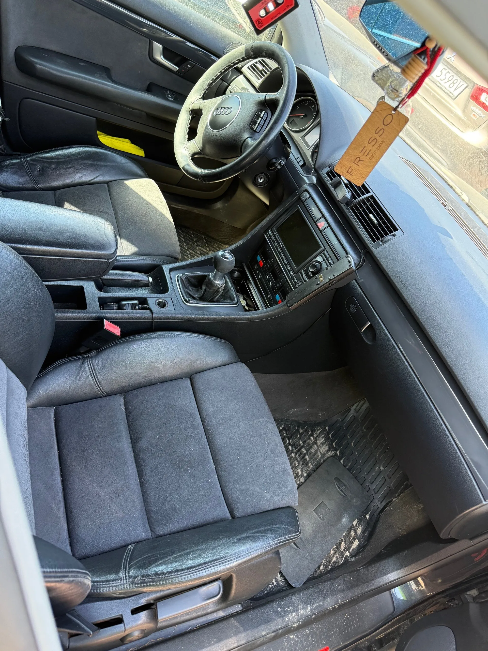 Wnętrze samochodu marki Audi z czarną skórzaną tapicerką. Widoczna kierownica z logo Audi, deska rozdzielcza z ekranem i przyciskami, dźwignia zmiany biegów oraz czarne dywaniki podłogowe. Na lusterku wstecznym wiszący drewniany znaczek z napisem FRESSO. Przez przednią szybę widoczny inny samochód z tablicą rejestracyjną 5598V. Wnętrze jest dobrze oświetlone, a siedzenia mają wyraźne ślady użytkowania.