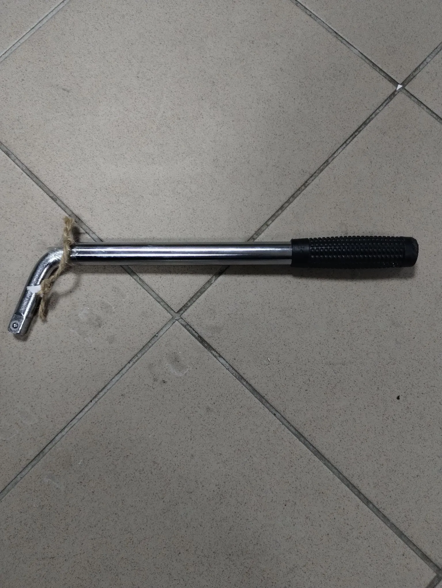 Metalowy klucz imbusowy o dużym rozmiarze, leżący poziomo na szarych, kwadratowych płytkach ceramicznych. Narzędzie posiada długą, chromowaną rączkę z czarną, gumową rączką na jednym końcu oraz zagięty koniec z sześciokątnym otworem na drugim. W miejscu zgięcia znajduje się kawałek brązowego sznurka.