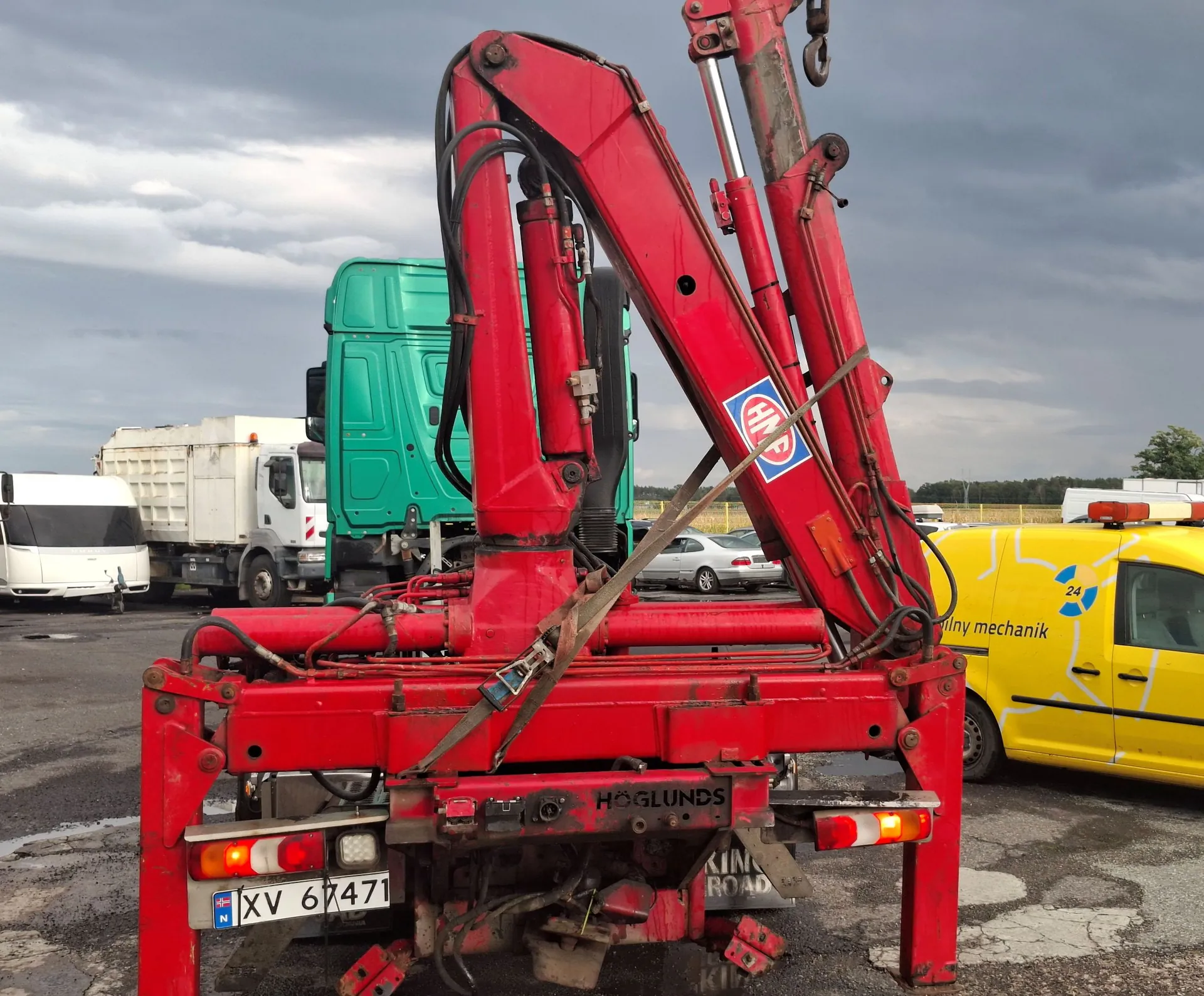 Czerwony żuraw hydrauliczny zamontowany na podwoziu pojazdu. W tle widoczna jest zielona ciężarówka, biała ciężarówka oraz żółty samochód dostawczy z napisem 'liny mechanik' i numerem '24'. Na czerwonej konstrukcji żurawia znajduje się logo oraz napis 'HÖGLUNDS'. Pojazd posiada tablicę rejestracyjną o numerze 'XV 67471' z oznaczeniem kraju 'N'. Całość znajduje się na asfaltowym placu pod zachmurzonym niebem.