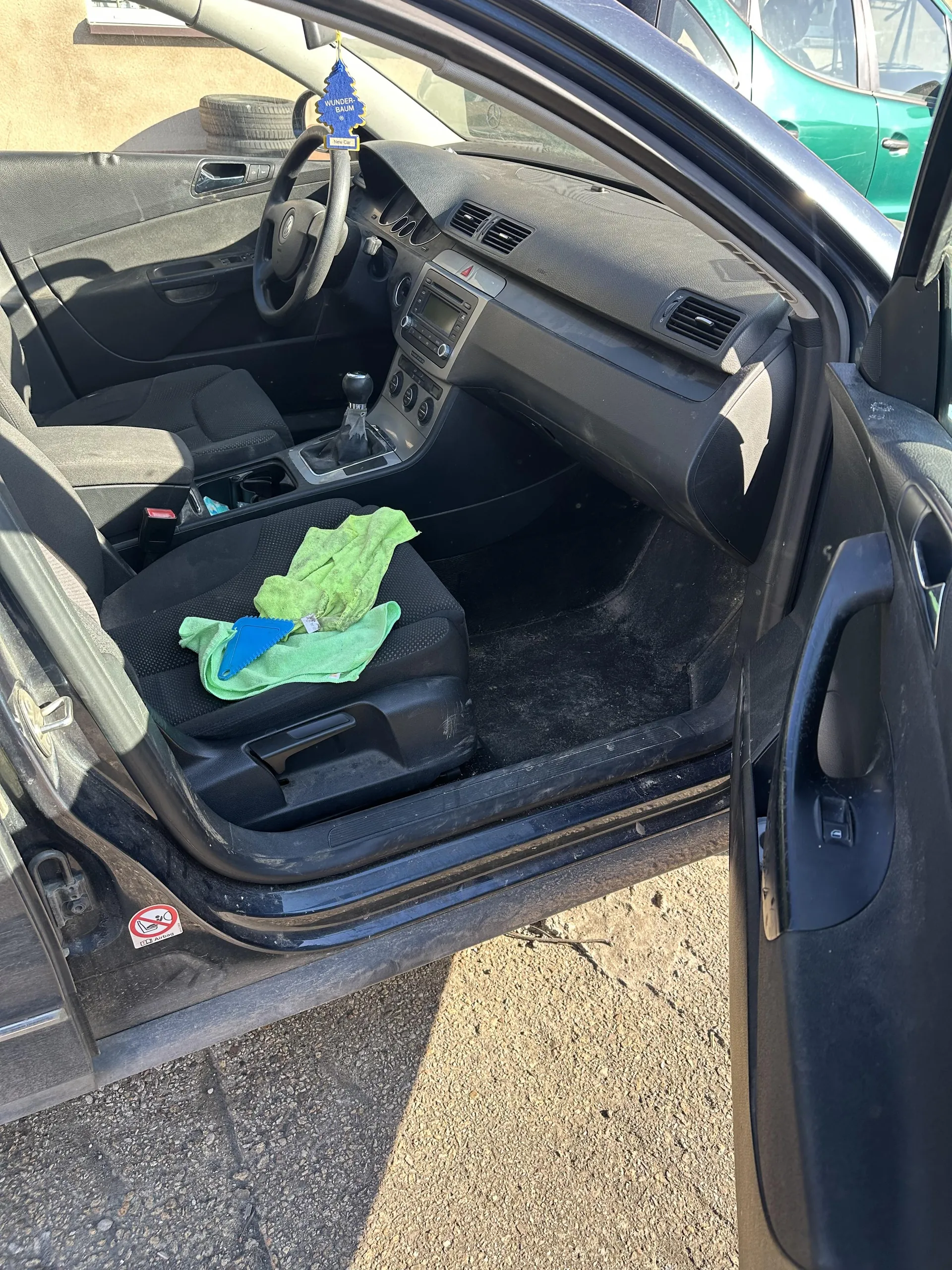 Wnętrze czarnego samochodu z otwartymi drzwiami od strony kierowcy. Widoczne czarne siedzenia, kierownica, deska rozdzielcza z radioodtwarzaczem i wentylacjami. Na siedzeniu pasażera leży zielona szmatka i niebieski przedmiot. Na lusterku wstecznym wiszący niebieski odświeżacz powietrza z napisem "WUNDER-BAUM". Poza samochodem widoczny zielony pojazd i opony na tle. Na drzwiach naklejka z zakazem palenia. Podłoga z asfaltu.