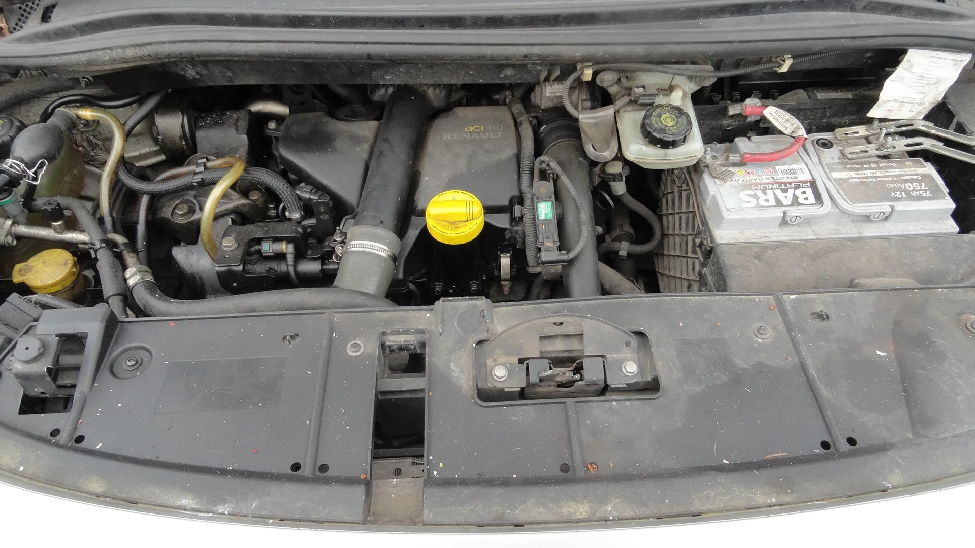 Otwarty maska samochodu z widocznym silnikiem marki Renault. Na silniku widoczne napisy 'dCi 110' oraz 'RENAULT'. W centralnej części znajduje się żółta pokrywka, prawdopodobnie do oleju. Po prawej stronie umieszczona jest bateria z napisami 'BARS', '750A 12V' oraz '750A'. Wokół silnika widoczne są przewody, węże i inne elementy mechaniczne. Na lewo od silnika znajduje się dodatkowa żółta pokrywka. Całość pokryta jest kurzem i śladami eksploatacji.