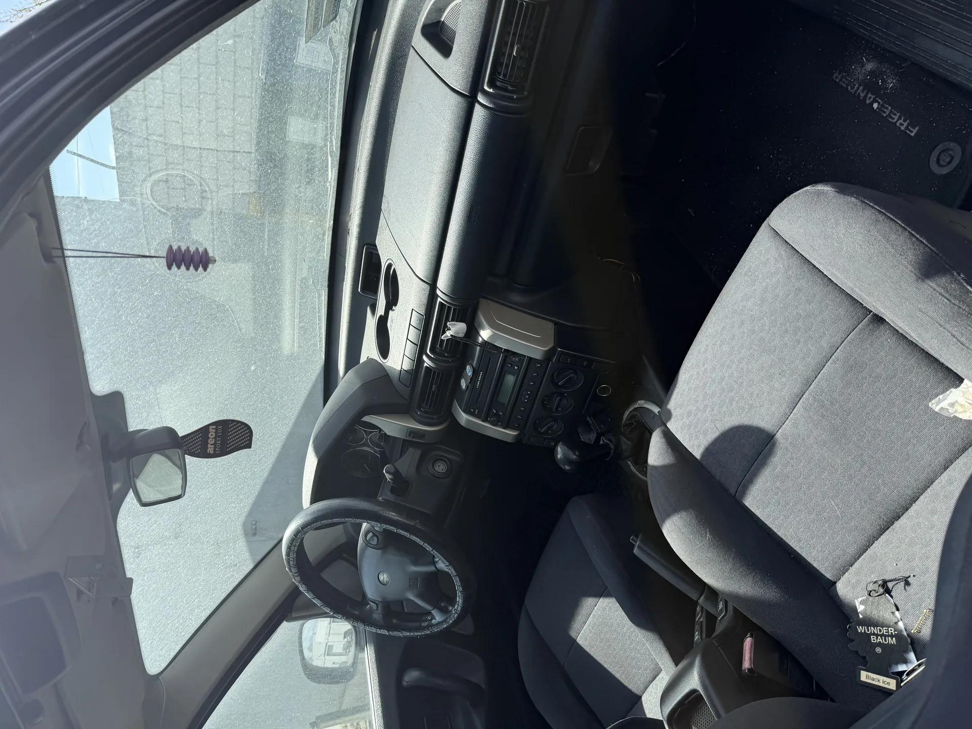 Wnętrze samochodu z widocznym kierownicą po lewej stronie, deska rozdzielcza z radioodtwarzaczem i przyciskami, szare siedzenia, wiszącym fioletowym odświeżaczem powietrza na lusterku wstecznym oraz etykietą z napisem WUNDER-BAUM i Black Ice na siedzeniu pasażera. Na dywaniku po stronie pasażera widoczny napis FREEDOM. Przez przednią szybę widać budynki i ulicę na zewnątrz.