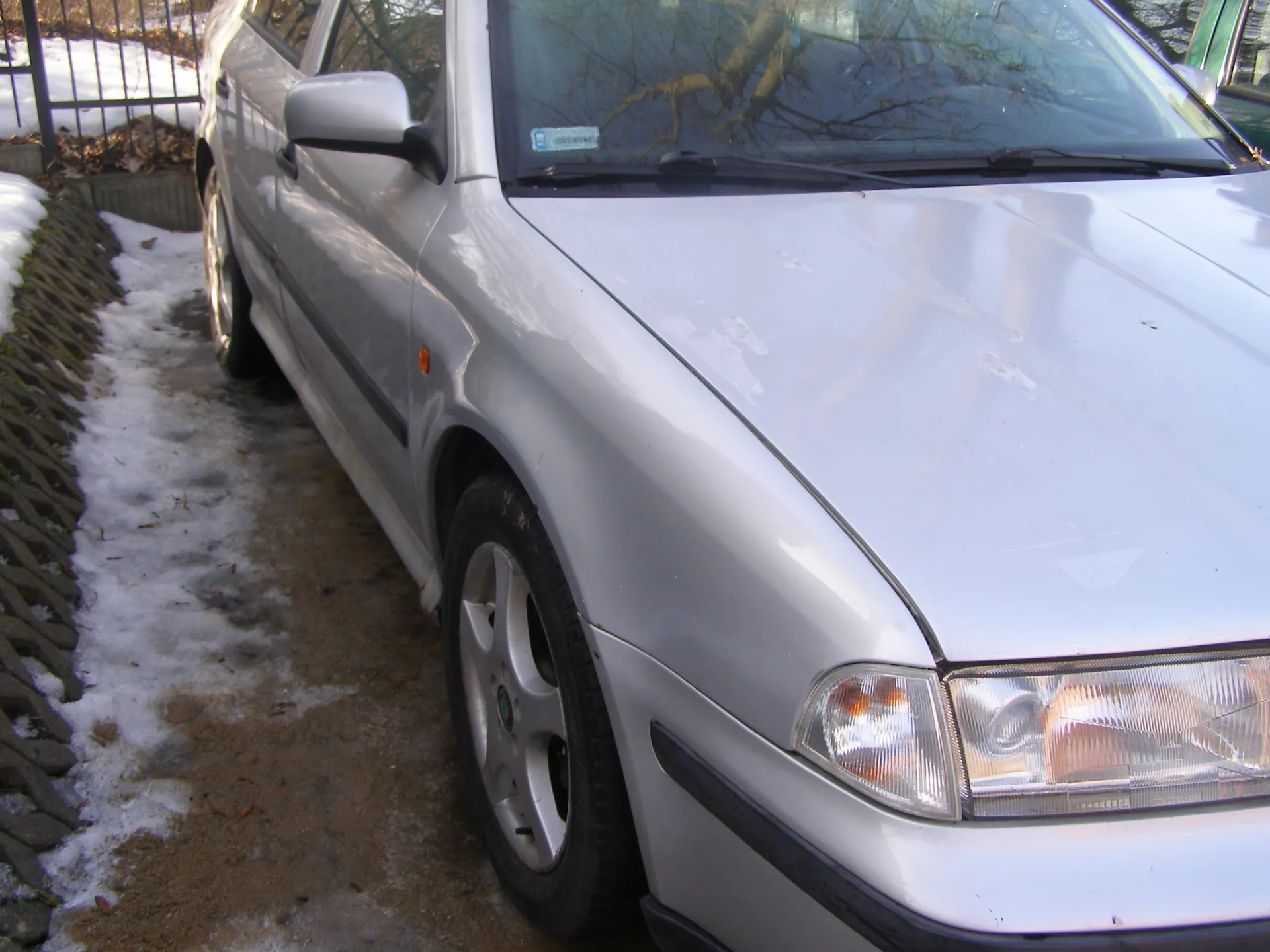Srebrny samochód osobowy zaparkowany na częściowo zasneżonym terenie. Widoczna lewa strona pojazdu, przednie koło, reflektory, lusterko boczne oraz naklejka na przedniej szybie. W tle metalowe ogrodzenie, śnieg, suche liście i drzewa bez liści. Na masce samochodu znajdują się drobne ślady. Teren pod pojazdem pokryty jest śniegiem i odsłoniętą ziemią.