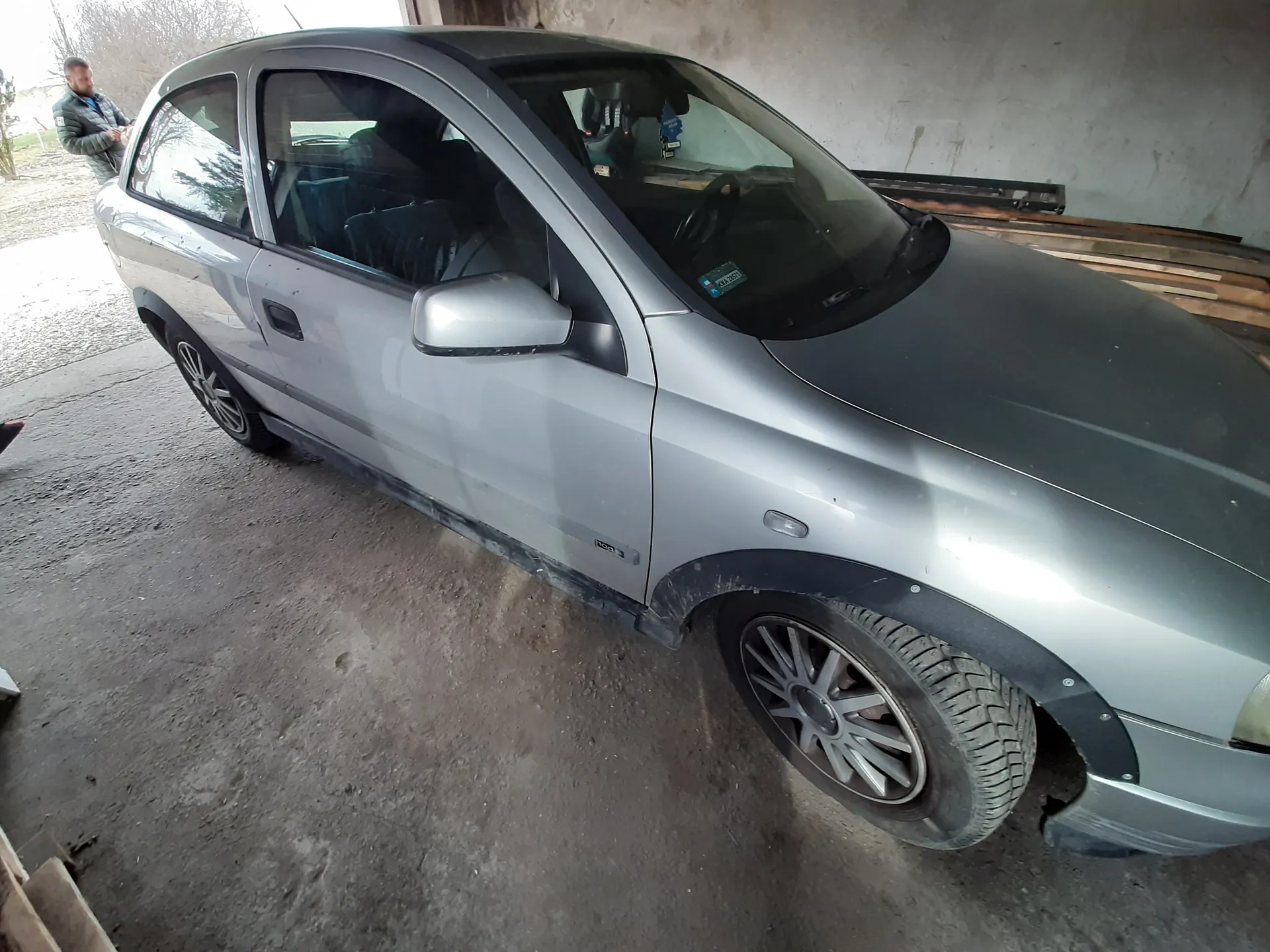 Srebrny trzydrzwiowy samochód osobowy zaparkowany w garażu z betonową podłogą i ścianami. Widoczne uszkodzenie przedniego zderzaka po prawej stronie. W tle mężczyzna w zielonej kurtce stojący blisko wejścia, a także drewniane deski oparte o ścianę. Na przedniej szybie naklejka z nieczytelnym tekstem. Opony z aluminiowymi felgami, podwozie częściowo zabrudzone.