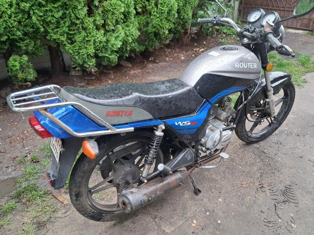 Niebiesko-srebrny motocykl marki Router, model WS 20, zaparkowany na błotnistym podłożu przed zielonym żywopłotem. Pojazd posiada srebrny bak z napisem ROUTER, czarne siedzenie, tylny bagażnik oraz czarne felgi. Na boku owiewki widnieje napis ROUTER oraz oznaczenie modelu WS 20.