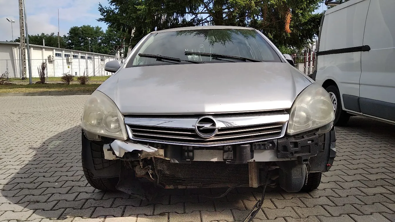 Srebrny samochód marki Opel z widocznymi poważnymi uszkodzeniami przedniego zderzaka, który jest częściowo oderwany, odsłaniając chłodnicę i elementy konstrukcyjne. Auto stoi na kostce brukowej obok białego vana. W tle widoczny jest biały budynek przemysłowy oraz zielone drzewa.