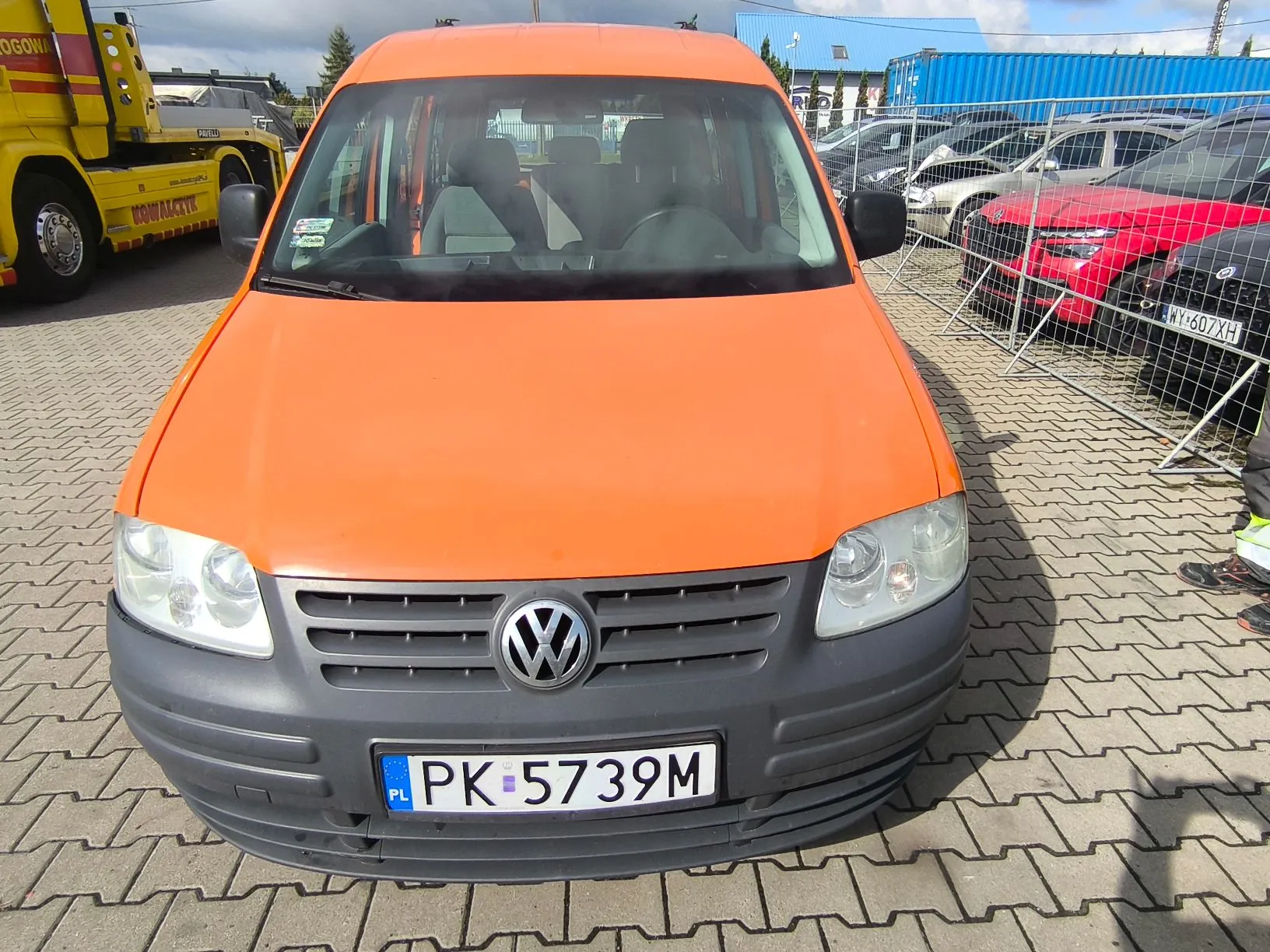 Pomarańczowy samochód dostawczy marki Volkswagen widziany od przodu, zaparkowany na kostce brukowej. Pojazd posiada polską tablicę rejestracyjną o numerze PK 5739M. W tle widoczne są inne samochody, w tym czerwony pojazd oraz żółty samochód specjalistyczny po lewej stronie.