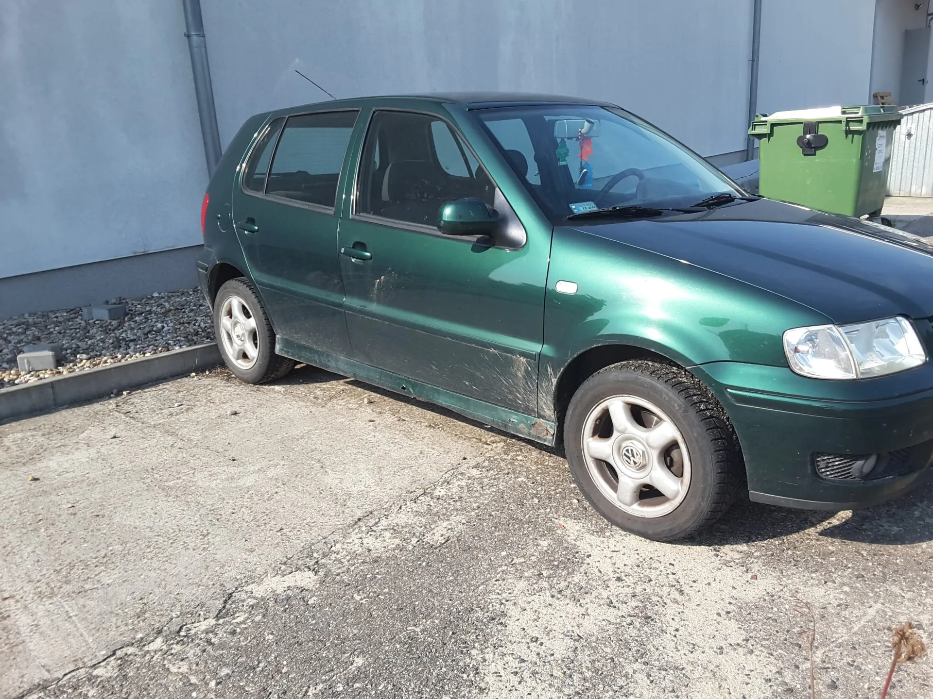 Zielony samochód osobowy marki Volkswagen Polo zaparkowany na betonowym podjeździe przed jasną ścianą budynku. Pojazd jest zabrudzony błotem w dolnych partiach nadwozia. W tle po prawej stronie widoczny jest duży, zielony kontener na odpady.