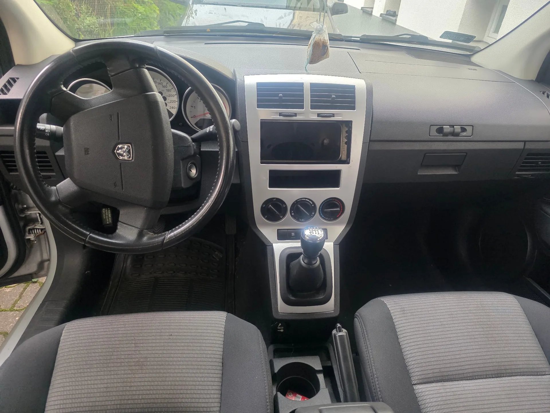 Wnętrze samochodu widziane z perspektywy przednich siedzeń. Widoczna jest czarna kierownica z logo marki Peugeot, deska rozdzielcza z licznikiem, centralna konsola z ekranem multimedialnym, pokrętłami klimatyzacji oraz lewarkiem zmiany biegów. W dolnej części zdjęcia znajdują się dwa szare materiałowe fotele z prążkowanym wzorem w centralnej części.