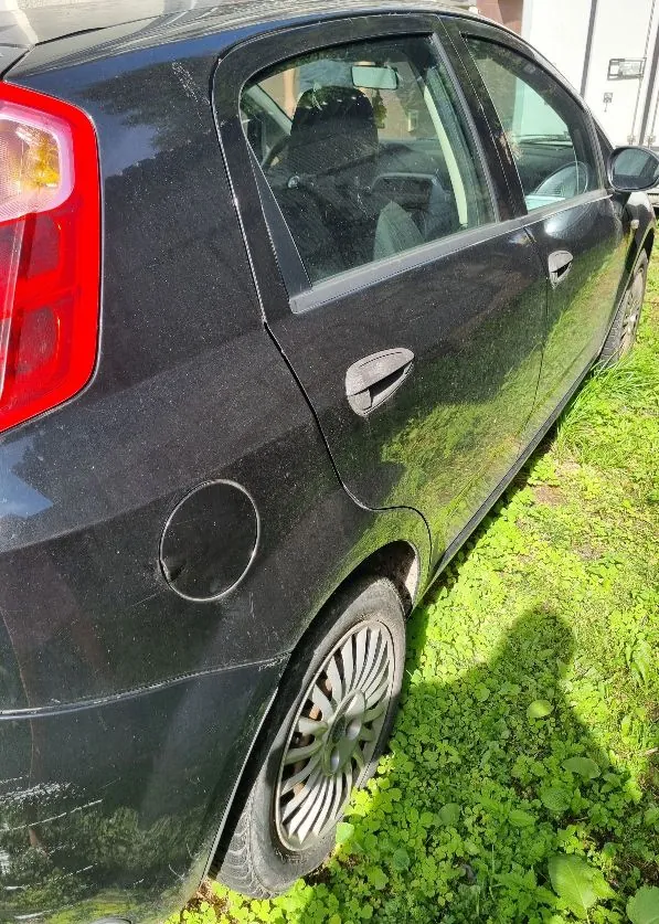 Zbliżenie na prawy bok czarnego samochodu osobowego. Widoczna jest tylna lampa, tylne i przednie drzwi z klamką, właz paliwa oraz tylne koło z wieloramienną felgą. Samochód stoi na trawie.